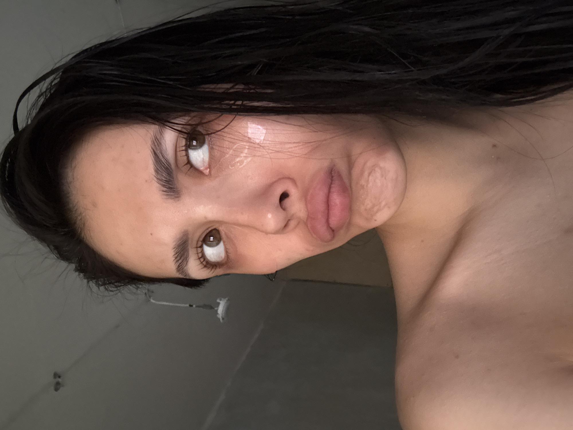 A close-up, rotated portrait shows a person with light skin, brown eyes, and dark, wet hair. The person is facing slightly upwards and towards the viewer, with their lips pursed. There is a clear, viscous substance or possibly moisture visible on the cheek. Part of the person's shoulder is visible in the lower right corner. The background appears to be a neutral grey color, possibly a tiled wall. There is no information available in the image about Votkinsk, Russia.