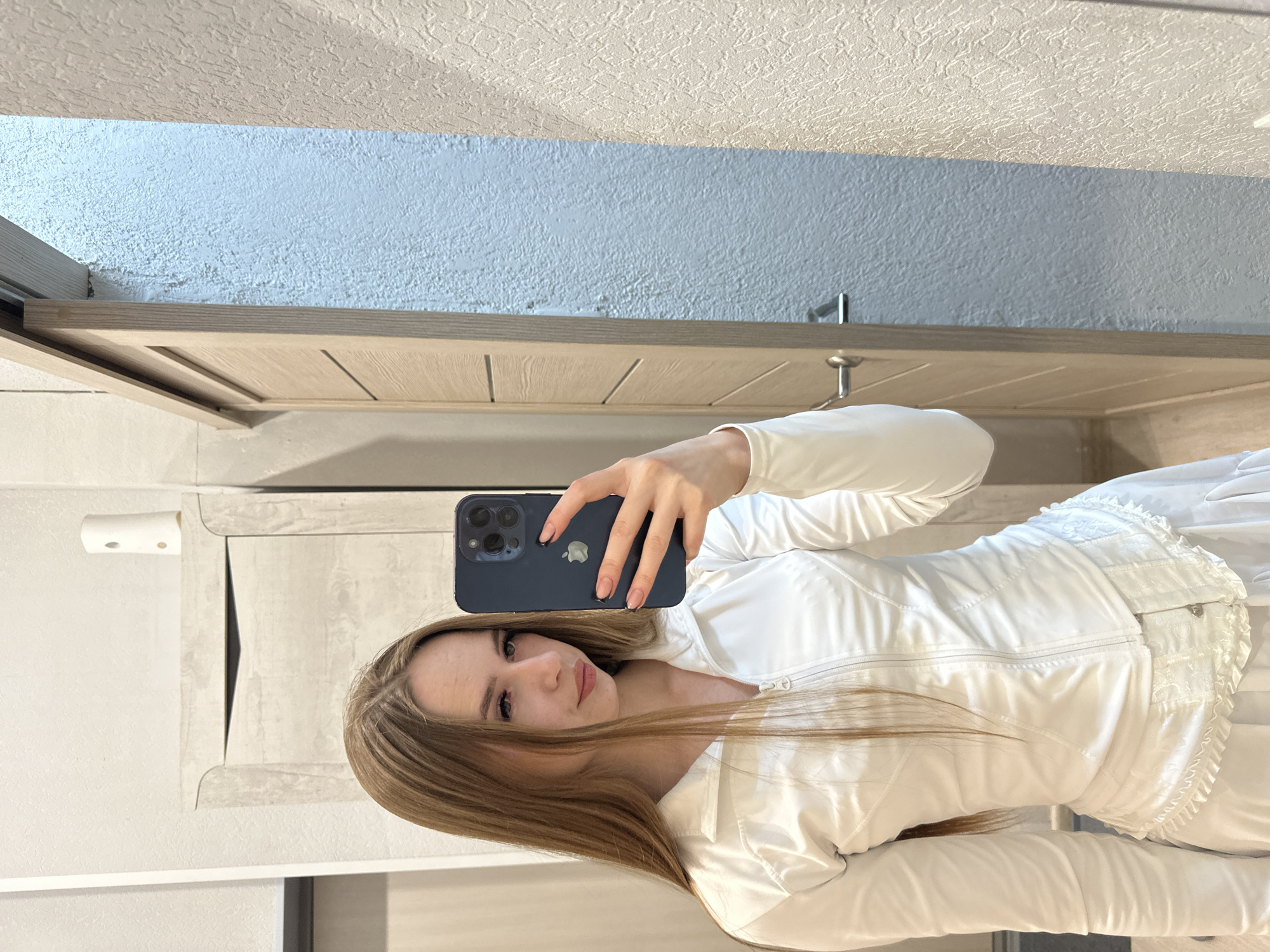 A woman is taking a selfie in what appears to be an indoor environment, potentially a bathroom. She is holding a dark blue smartphone in her right hand, obscuring part of her face. She has light brown hair and is wearing a white zippered jacket and a white outfit with frills along the side. The background shows a light wood-grained shelf or cabinet, a light blue wall with a textured surface, and a white ceiling. The location is unknown.