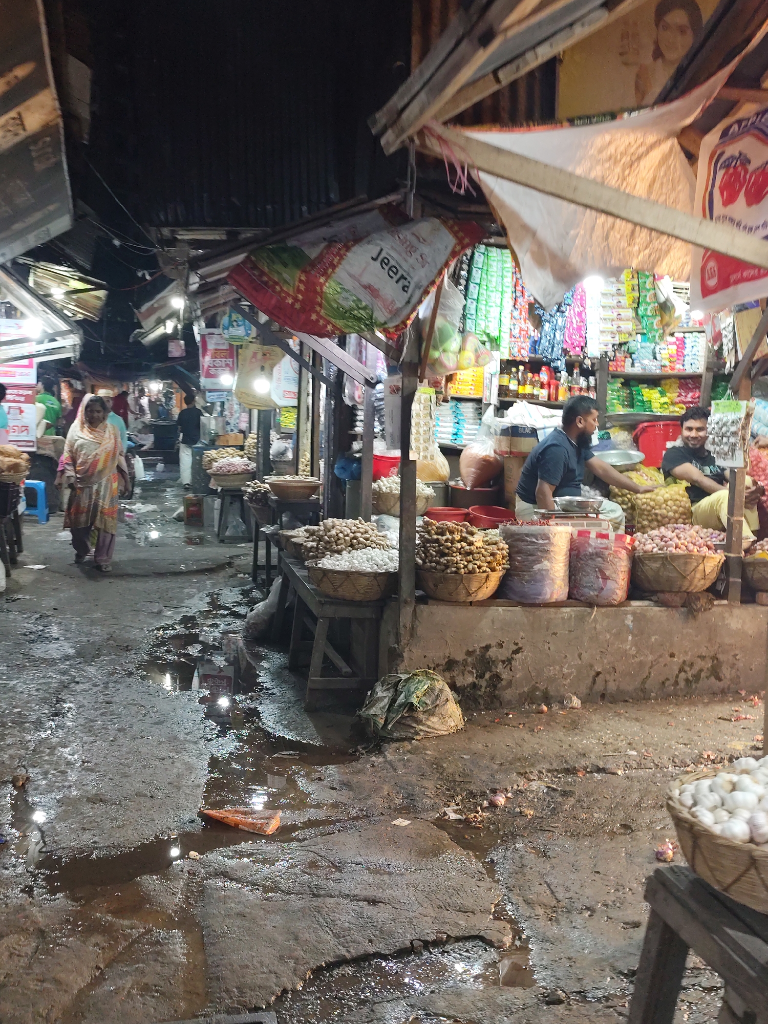 The image depicts a bustling marketplace in Dhaka, Bangladesh. Two men are seated behind a display of produce, including ginger, onions, and potatoes, arranged in baskets and bags. The marketplace appears to be crowded, with people walking along a pathway. The ground is wet and uneven, with puddles of water reflecting the ambient light. Overhead, various signs and advertisements are displayed. The market structure is a mix of temporary and semi-permanent structures, with tarps and awnings providing shelter. The overall atmosphere is lively and crowded, indicative of a typical market scene in Dhaka.