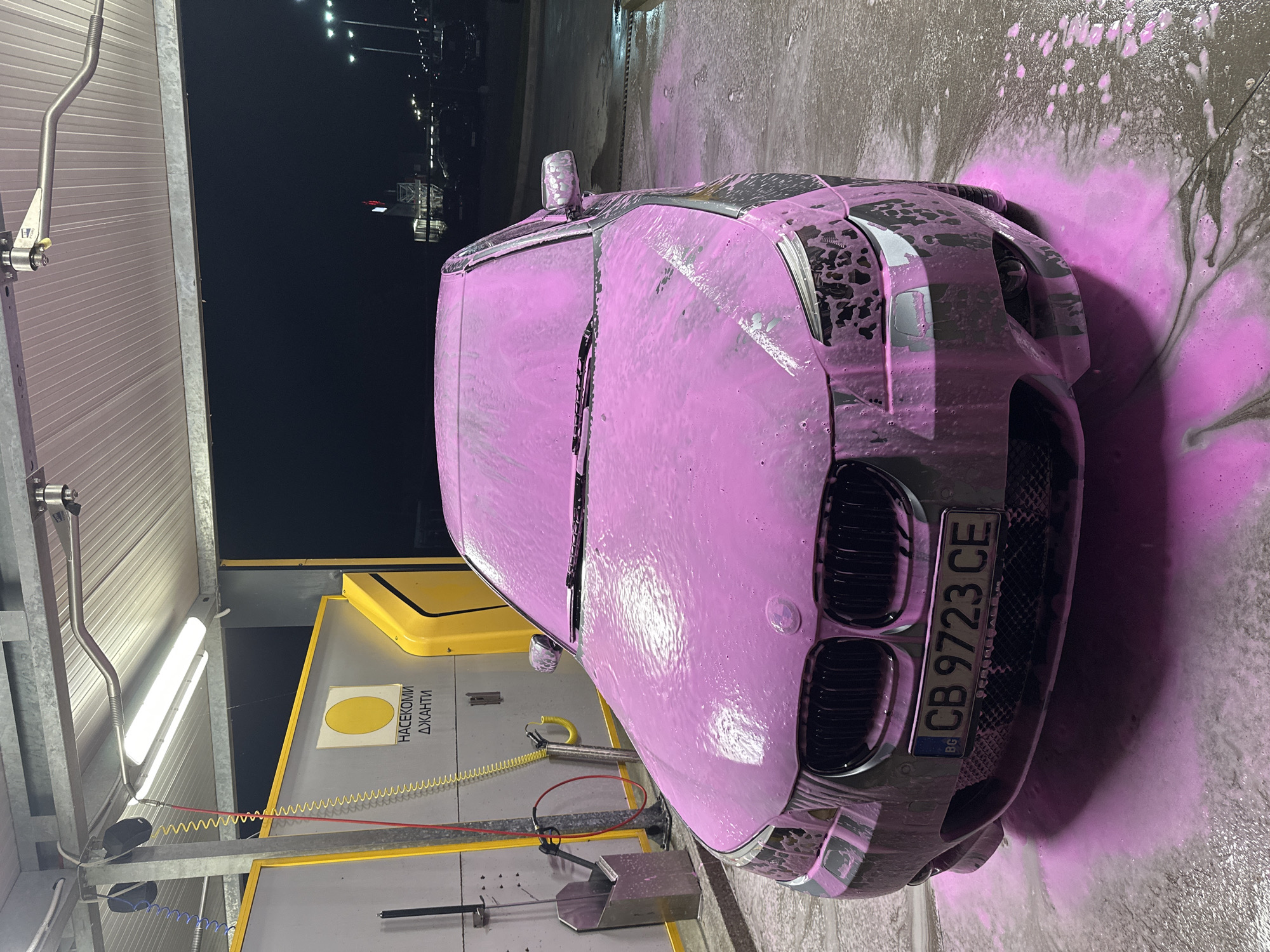 The image shows a car being washed with pink foam at a self-service car wash station. The vehicle has a Bulgarian license plate "CB 9723 CE". The car wash station has a yellow panel with the words "НАСЕКОМИ ДЖАНТИ" printed in Cyrillic. The location is likely in Plovdiv, Bulgaria, based on the license plate. The sky is dark, suggesting it is night or early morning. There are lights visible in the background.