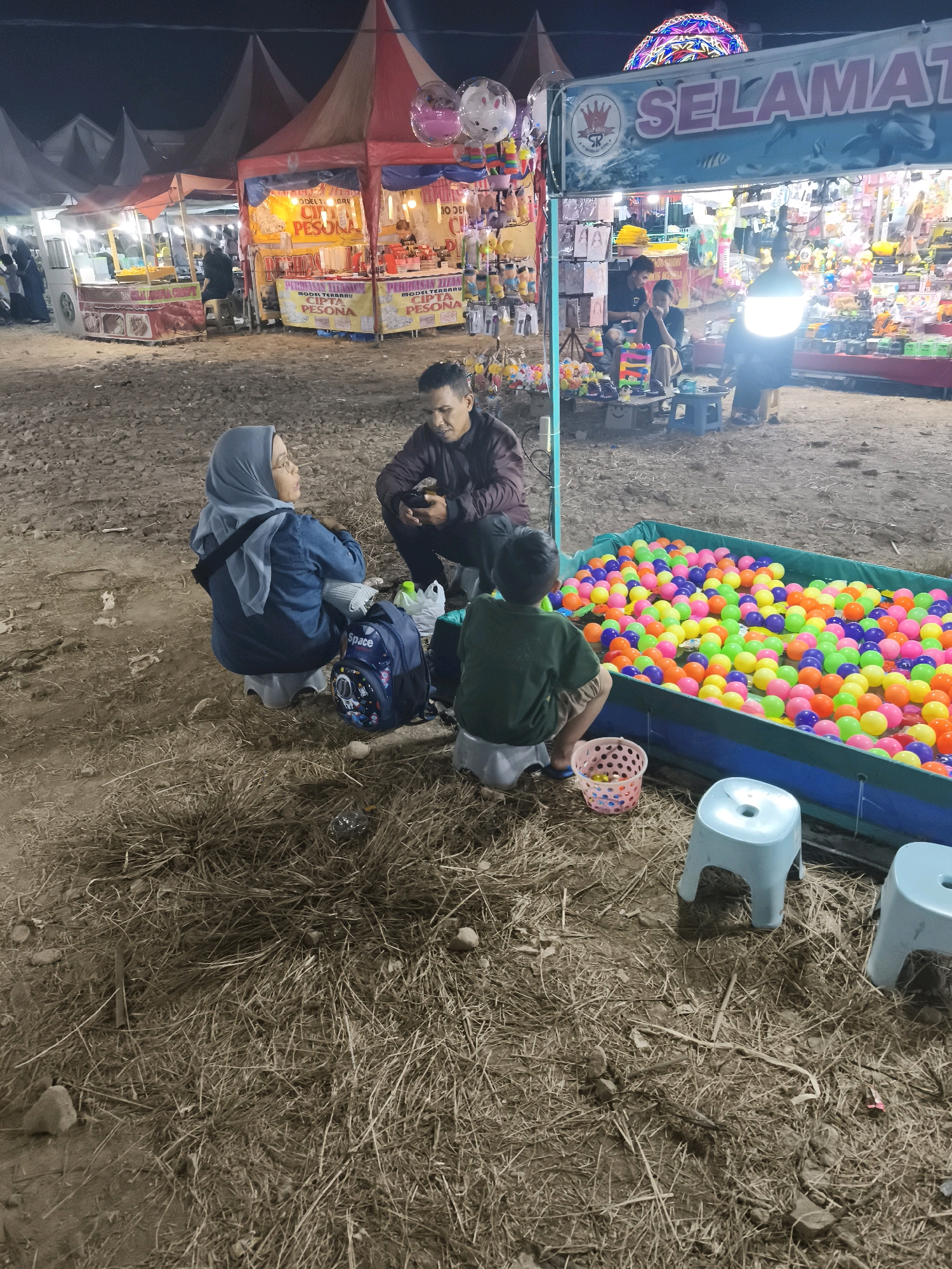 This image captures a vibrant night scene at a carnival or fair in Palembang, Indonesia. In the foreground, three individuals are seated. A woman wearing a blue hijab and denim jacket sits on a small stool, looking to her left. Next to her, a man in a dark jacket is also seated, holding a smartphone. A young boy, wearing a green t-shirt and khaki shorts, sits on another stool facing a large, blue-lined ball pit filled with numerous brightly colored plastic balls. A pink basket with a few yellow balls sits beside him.
In the background, several market stalls are visible under brightly lit tents. One stall features a large banner with the word "SELAMAT" in prominent lettering. Other stalls display various goods, including toys and merchandise, illuminated by strings of lights. The ground is covered with dry grass and scattered debris, suggesting an outdoor, somewhat unkempt location. The overall atmosphere is festive and lively, typical of a community event.