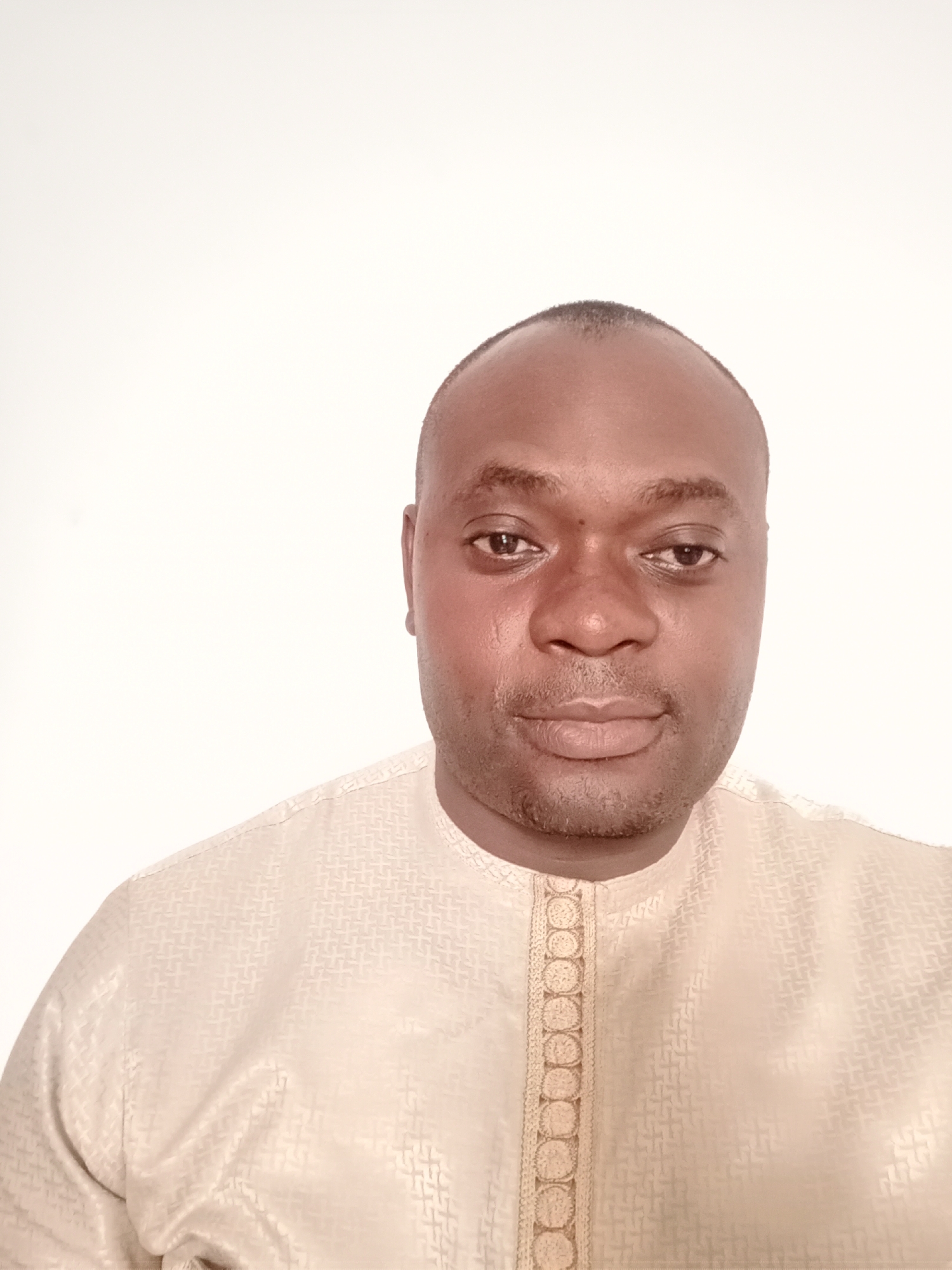 A close-up studio portrait of a dark-skinned man with a shaved head and a trimmed goatee, wearing a light beige traditional African garment, likely a Hausa Buba, is set against a stark white background. The details of the garment near the collar show a pattern design and decorative trim. The man looks directly at the camera with a neutral expression. There is no indication that the image was taken in Gwarinpa, Nigeria, or any other specific location. The description is based solely on the visual information in the provided image.