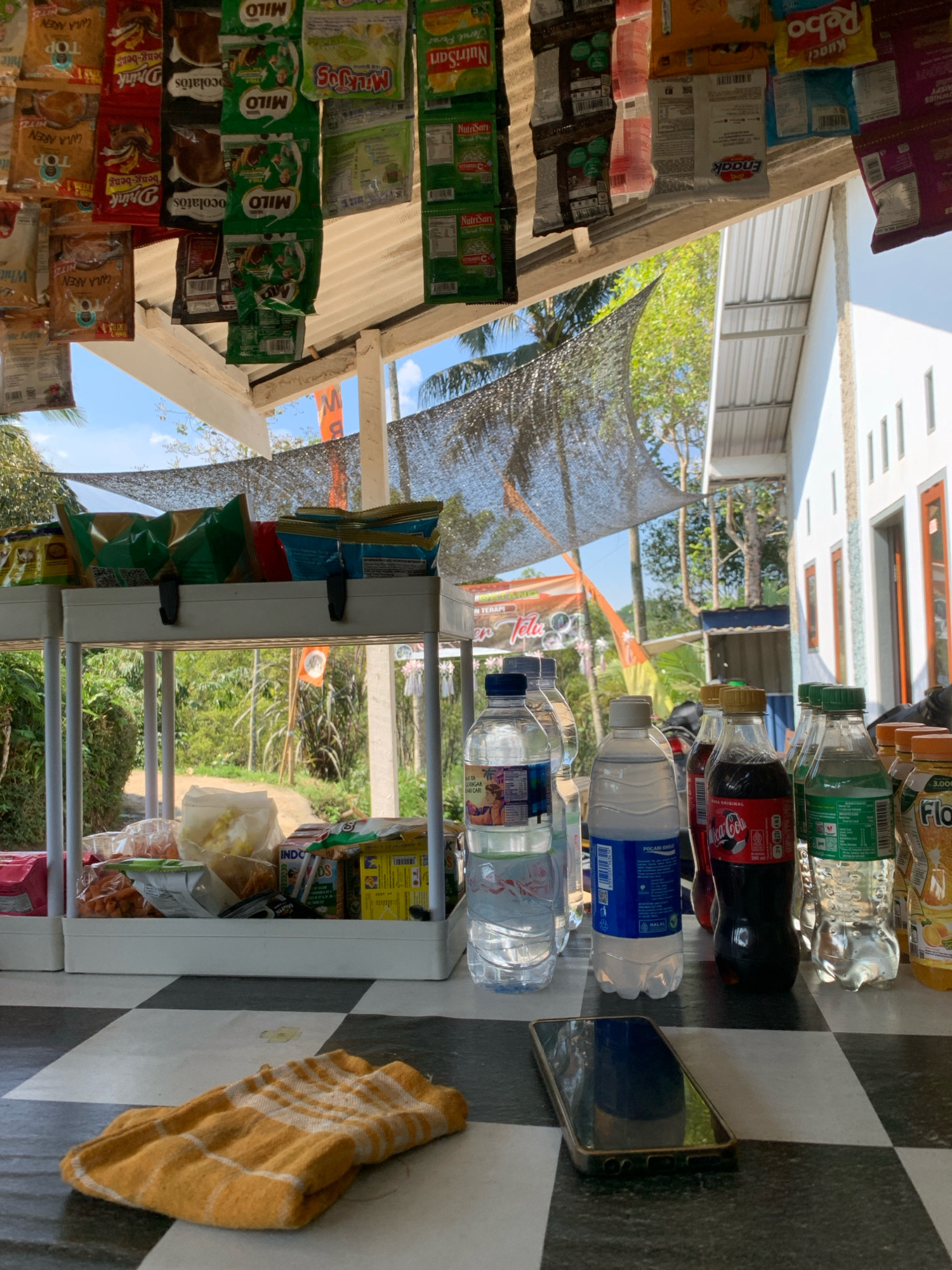 The image depicts an outdoor stall in Pronojiwo, Indonesia, likely at a roadside market or warung. The stall is set up under a sloped, translucent roof, with various packaged snacks and drinks hanging from the ceiling. In the foreground, a black and white checkered surface serves as a table or counter, on which several plastic bottles of beverages are arranged. These include what appear to be water bottles, a bottle of Coca-Cola, and a bottle of orange-flavored drink. A folded yellow and white striped cloth is also present on the checkered surface, as is a smartphone. Behind the bottles, a white two-tiered shelf holds more packaged goods, including bags of snacks and boxes. The background reveals a lush green natural environment with trees and foliage, suggesting a rural or semi-rural setting. The bright sunlight indicates it is daytime. The overall scene suggests a small, local business operating in a tropical climate.