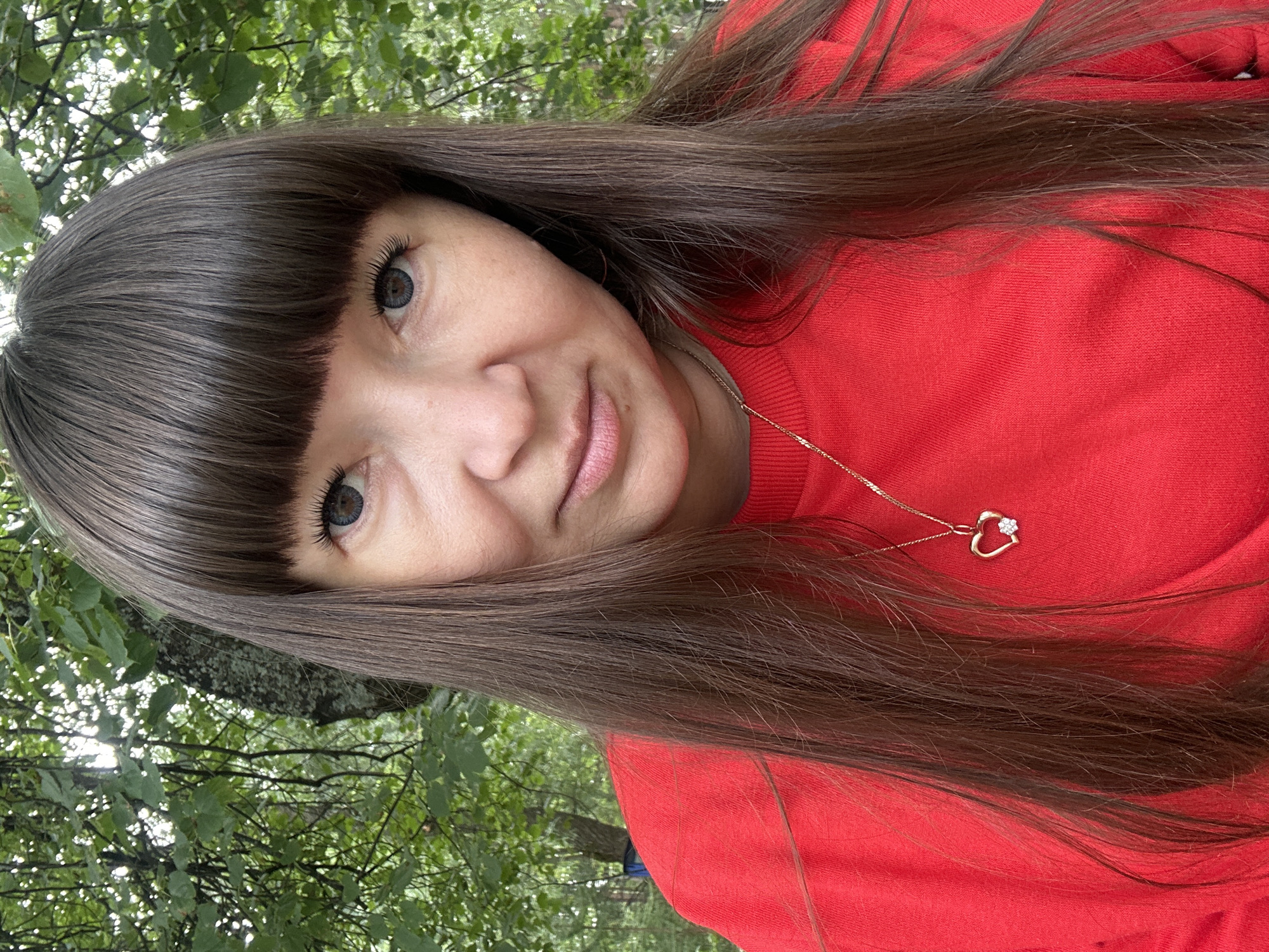 A medium shot photograph captures a woman with long, dark brown hair and bangs, wearing a bright red sweatshirt and a gold heart-shaped pendant with a small white stone. She is outdoors, with a blurred background of green trees and foliage, suggesting a natural or park-like setting. The lighting is soft and diffused, indicating an overcast day. There is no identifiable information in the image to suggest a specific location within the city of Chaikovsky, Russia.