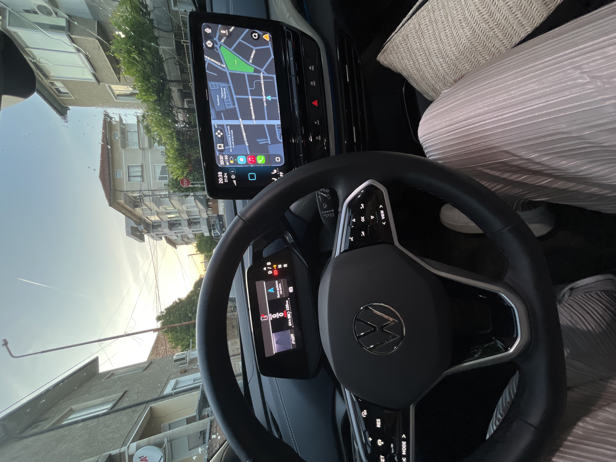 The image shows the interior of a Volkswagen car, focusing on the dashboard, steering wheel, and infotainment system. The navigation system displays a map with recognizable streets and landmarks in the city of Unknown, Unknown, including Kaufland, ул. Стара планина, ул. Стоян Бъчваров, парк, ул. Цар Симеон Велики, and ул. Искър. The time on the display indicates 20:38, and the temperature is 21.0°C. The instrument cluster shows the car is in park with a battery level of 64% and an estimated range of 230km. The steering wheel features the Volkswagen logo and control buttons. A person wearing light-colored clothing is partially visible in the passenger seat. The exterior view through the windshield reveals buildings and trees.