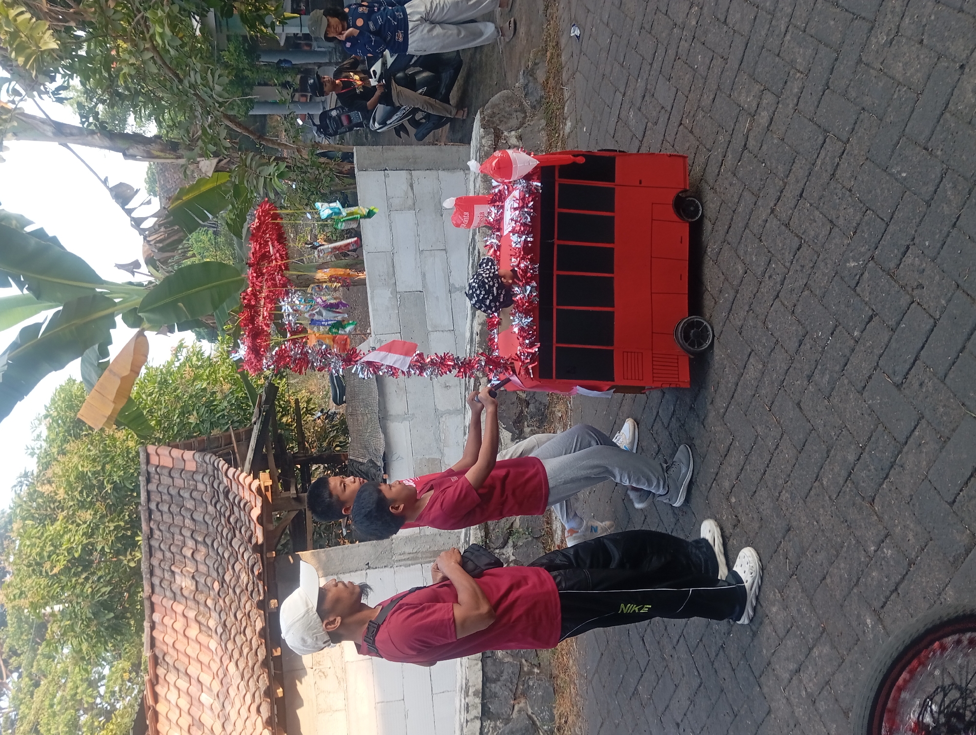 In Jogosari, Indonesia, three individuals are engaged in a parade. A man wearing a white cap, red shirt, and black pants with white sneakers is walking on the left. Next to him, two boys in red shirts and grey pants are pushing a red bus-shaped float. The float is decorated with red and silver tinsel, small flags, and balloons. In the background, other people are present, along with motorcycles and buildings with tiled roofs. The scene takes place on a paved street lined with trees and a white brick wall.