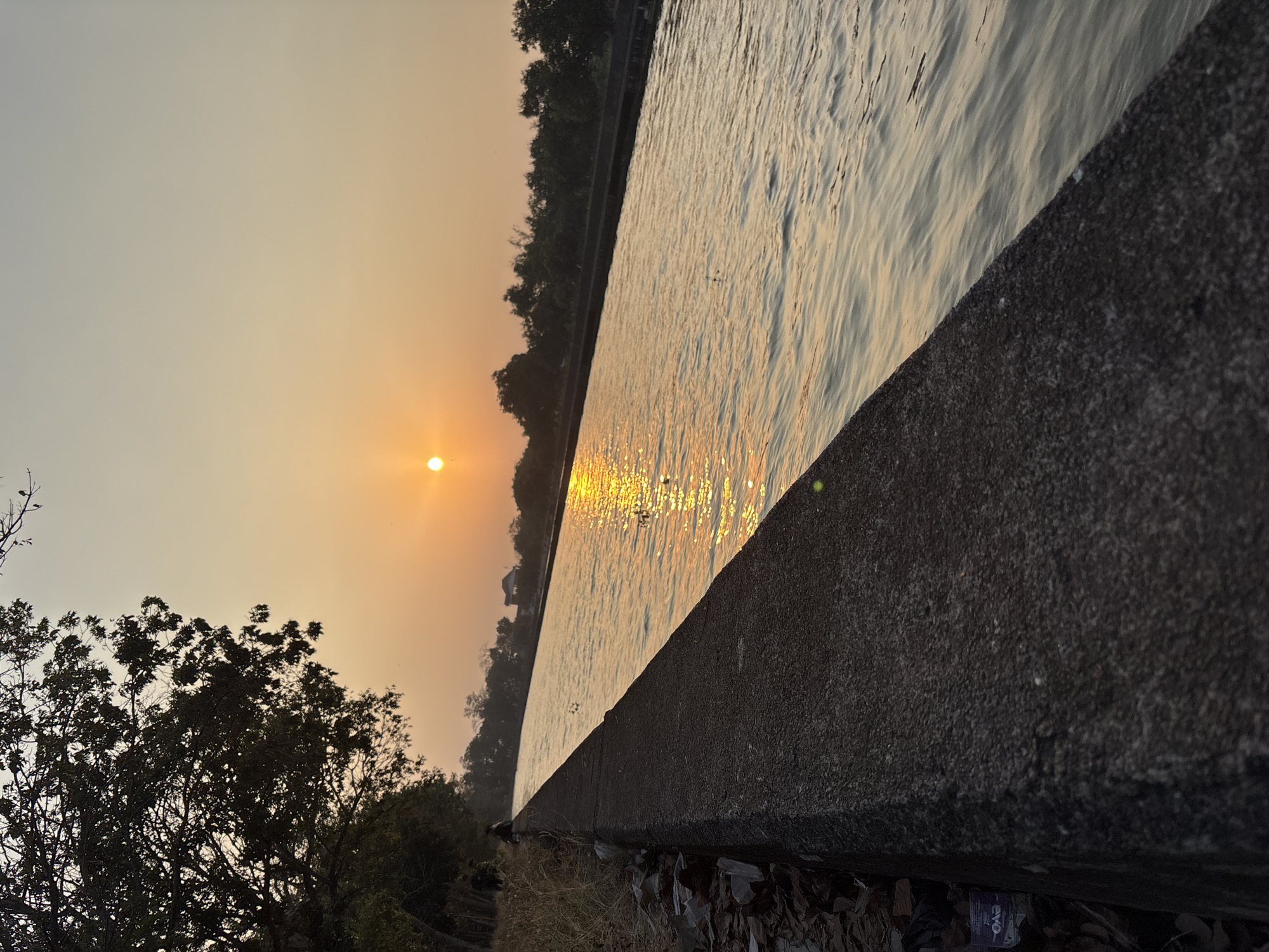 A wide, grey concrete embankment slopes diagonally from the lower left to the upper right of the frame, bordering a calm, wide canal filled with water. The canal reflects the warm, golden light of a setting sun, which appears as a bright, orange orb positioned in the upper center of the sky. Trees with dark foliage line the far bank of the canal, creating a silhouetted backdrop against the hazy, orange-tinted sky. A few scattered, indistinct figures are visible along the embankment and on the far bank. The foreground of the embankment is littered with dried leaves and some debris, including a small, faded purple package. The overall scene captures a tranquil, late afternoon or early evening atmosphere.