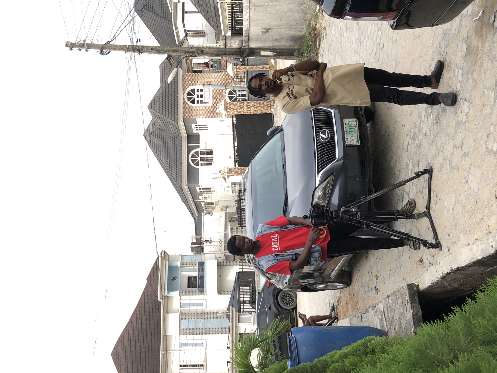 This image captures a daytime scene on what appears to be a residential street in Lagos, Nigeria. Two men are featured alongside a dark grey Lexus SUV.
On the left, a man wearing a red t-shirt with "CATAL" printed on it, a denim vest, and black pants is operating or setting up a professional video camera mounted on a black tripod. He is focused on the camera, which is positioned in front of the Lexus SUV. The SUV's license plate, "LAGOS EKY-391FL," is clearly visible.
To the right of the SUV, a second man stands, wearing a light brown traditional African top, black pants, and dark shoes. He has dreadlocks or braids and is looking towards the camera with a neutral to slightly smiling expression, his right hand resting near his chin.
The background reveals several multi-story houses with distinct architectural features, including tiled roofs, arched windows, and patterned facades, consistent with modern residential buildings. A wooden utility pole with numerous wires extends upwards on the far left. The ground is paved with interlocking stones, and a drainage channel runs along the right side. In the lower-left foreground, green foliage and two large blue barrels are partially visible. The scene is brightly lit, indicating it is daytime, and suggests an outdoor video production or photoshoot is taking place.