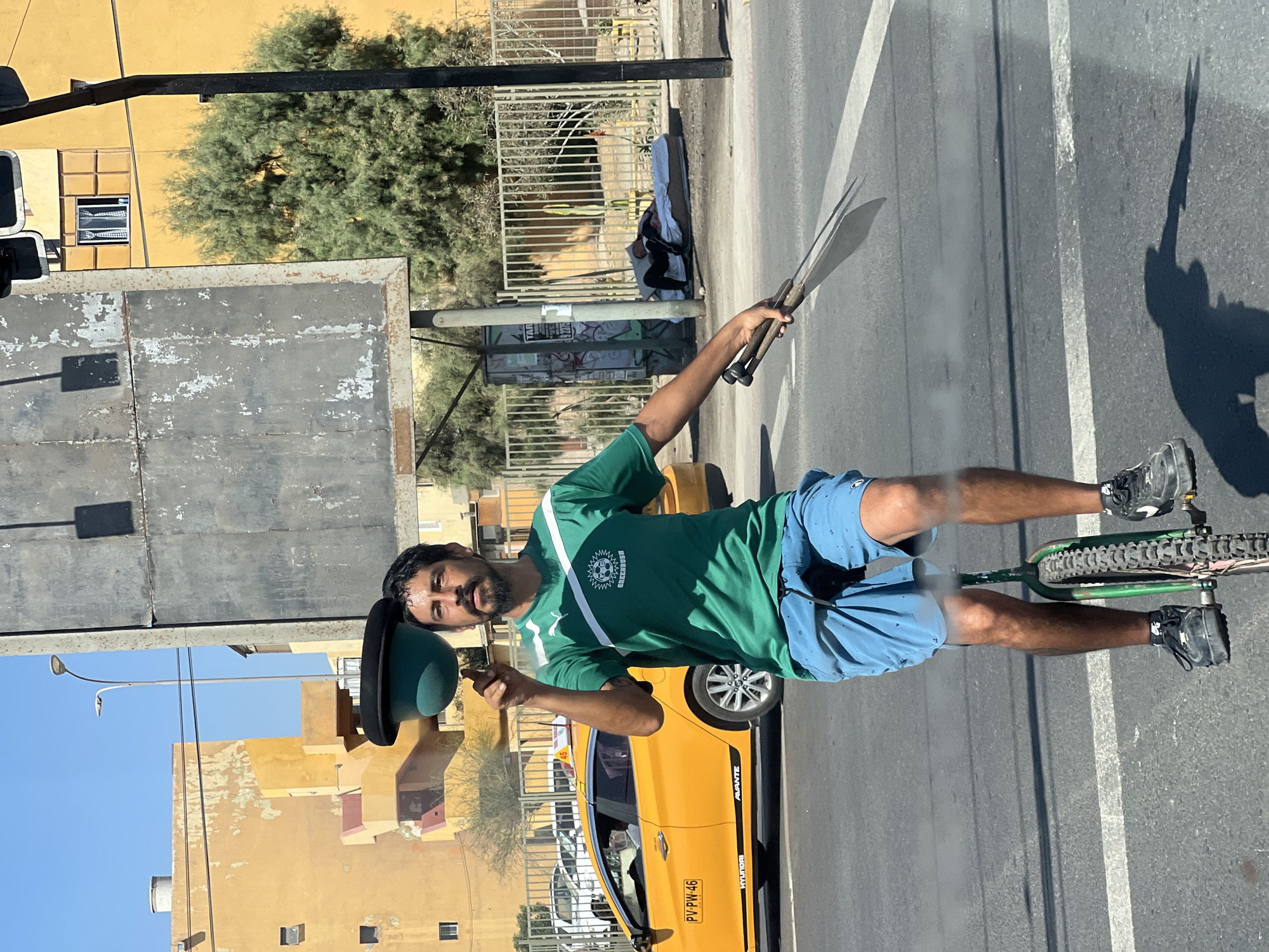 Arica, Chile, street scene. A man on a green unicycle holds two long machetes in his right hand and a green bowler hat in his left. He wears a green athletic shirt with a white diagonal stripe and blue shorts. A yellow taxi cab is visible behind him. Buildings and trees are in the background. A person sleeps on a mattress next to a bus stop.