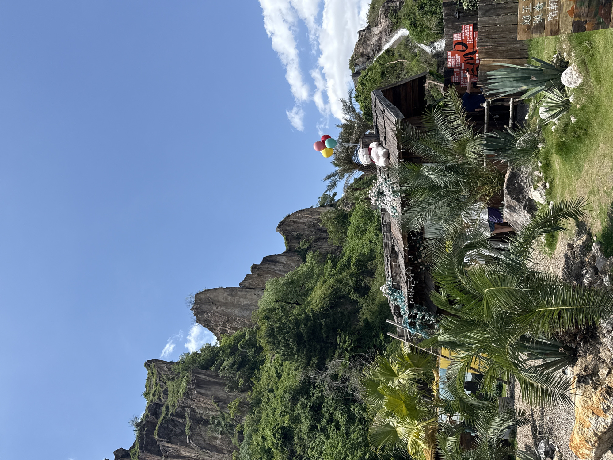 The image shows a view of a building next to a cliff in Keqiao District, China. The building is made of wood and has a thatched roof. There are several palm trees around the building. A waterfall can be seen in the background. The sky is blue and there are a few clouds. A bunch of colorful balloons are visible on the roof of the building next to a few plush toys. People are visible on the roof of the building.