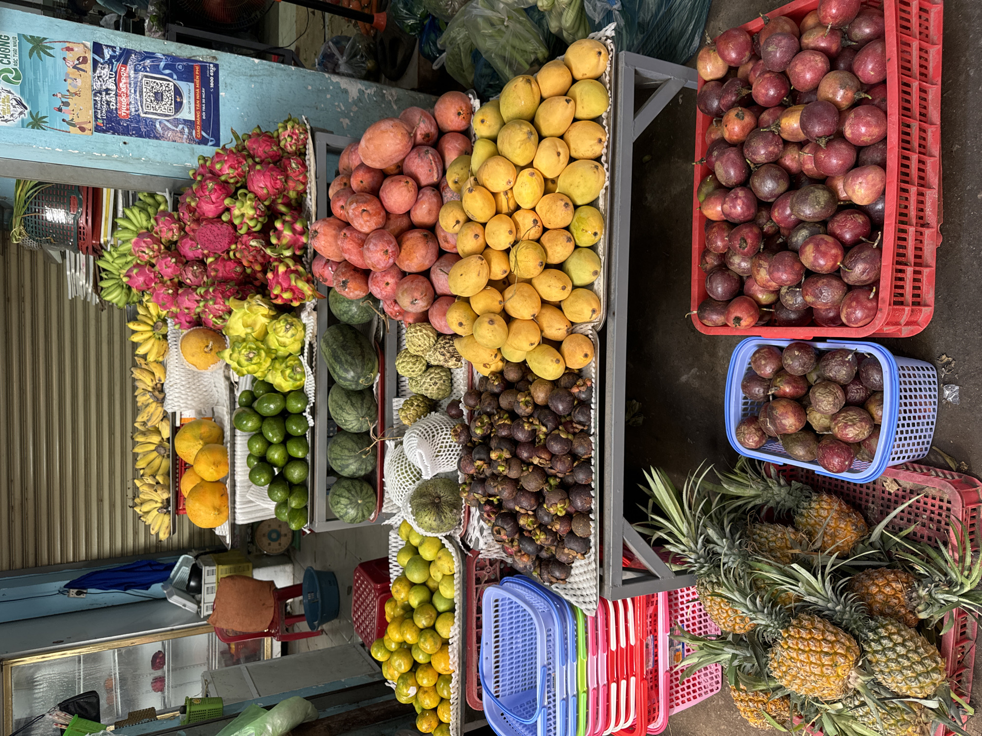 The image depicts a vibrant fruit stand in Mũi Né, Vietnam. Bananas, dragon fruit, pink and yellow mangoes, passion fruit, custard apples, mangosteen, oranges, watermelons, avocados, and pineapples are densely arranged on a tiered metal shelf and in plastic baskets. A blue advertisement with a QR code is affixed to a nearby surface. The setting is a covered market area, indicated by a partially visible shutter and nearby infrastructure.