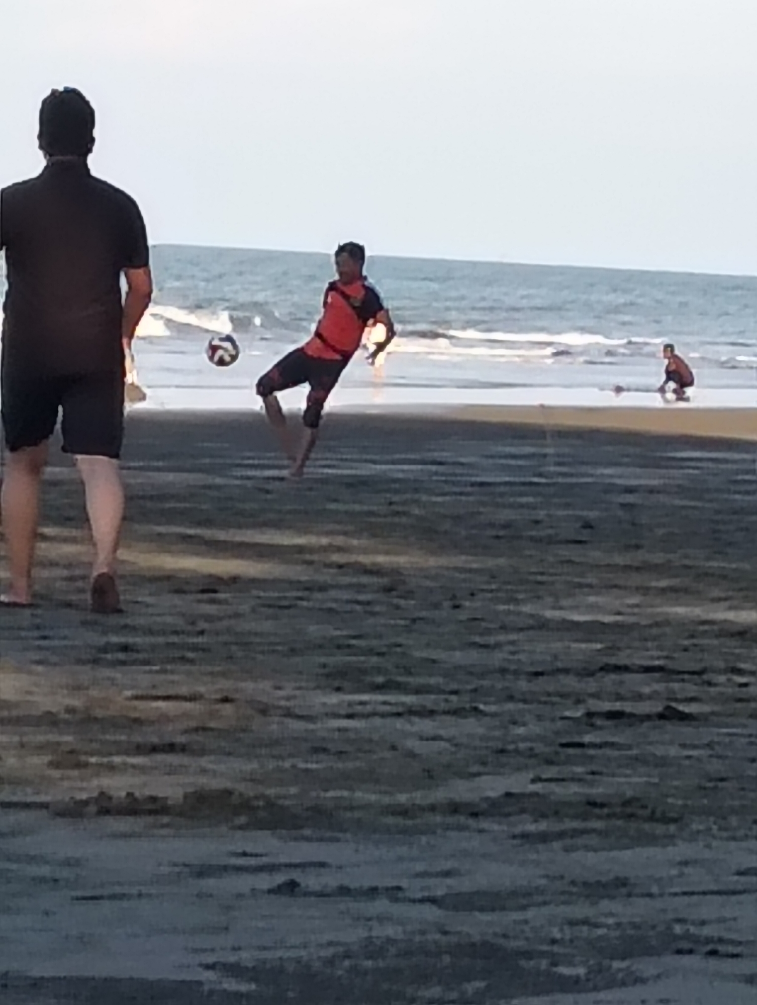 The image depicts a beach scene in Peukan Sot, Indonesia, with several individuals engaged in recreational activities. In the foreground, a person stands facing away from the camera, wearing a black t-shirt and dark shorts. To their right, another person is captured mid-action, kicking a soccer ball. This individual is wearing an orange and black striped shirt and dark pants. Further in the background, a third person is seen crouching on the sand near the water's edge, facing the ocean. The environment is a sandy beach with gentle waves lapping the shore. The sky is overcast, and the overall lighting suggests it may be late afternoon or early morning.