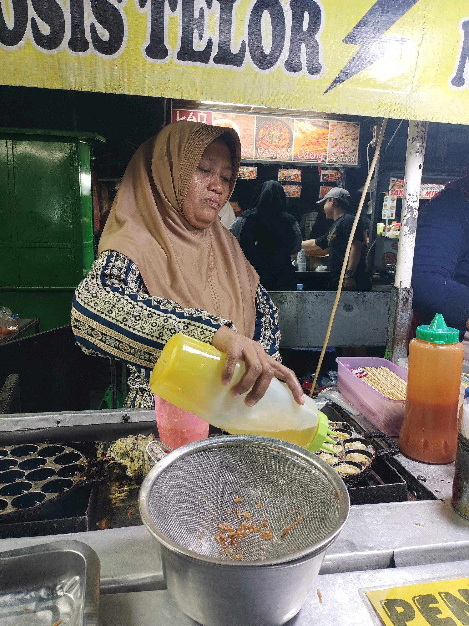In Surabaya, Indonesia, a woman wearing a light brown hijab and a patterned blue and white shirt is seen preparing food. She is holding a large yellow squeeze bottle, from which she is pouring a liquid into a batter-filled cooking mold, likely for a street food snack. In front of the cooking mold is a metal sieve containing some fried ingredients. Next to the sieve is a pink container filled with wooden skewers. To the right of that, an orange squeeze bottle with a green cap is visible. The scene appears to be at a street food stall, with a yellow banner featuring large black lettering at the top. Behind the woman, other people are visible, and there are posters advertising various food items on display. The lighting suggests it is evening or nighttime.