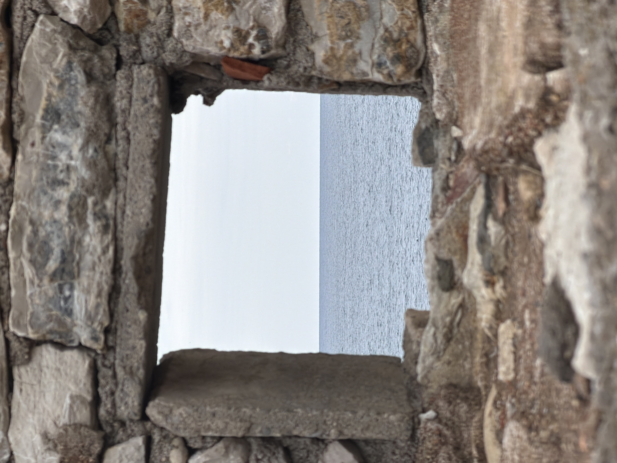 A view of the sea through a stone window in Petrovac centar, Montenegro. The window is made of grey stone, with rough textures and visible wear. The left side of the frame is composed of large, irregular stones, while the right side shows a more fragmented, weathered appearance. The window opening reveals a horizontal division, with a pale, nearly white sky above, and the grey, rippled surface of the sea below. The horizon line is slightly tilted. The scene is still.