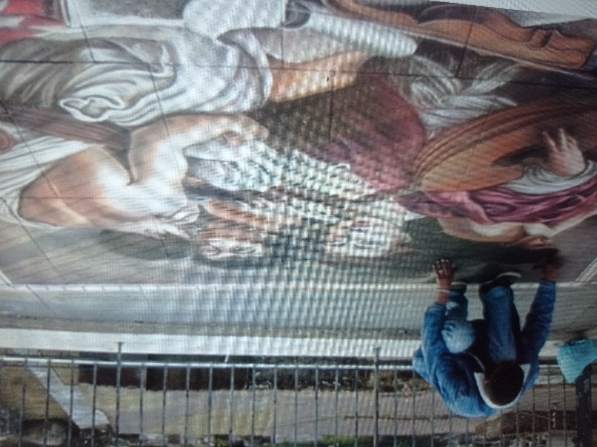 The image depicts a street art chalk drawing of two figures lying down, one holding a stringed instrument. A person, dressed in blue, is kneeling on the ground to the left of the drawing, appearing to be working on it. The scene is set on a sidewalk with a metal fence and a street in the background. The chalk drawing is detailed and appears to be a realistic depiction of classical figures. The location is identified as Mukachevo, Ukraine, suggesting this is a public art installation. The perspective is from above, looking down on the scene. The lighting is natural, indicating daytime.