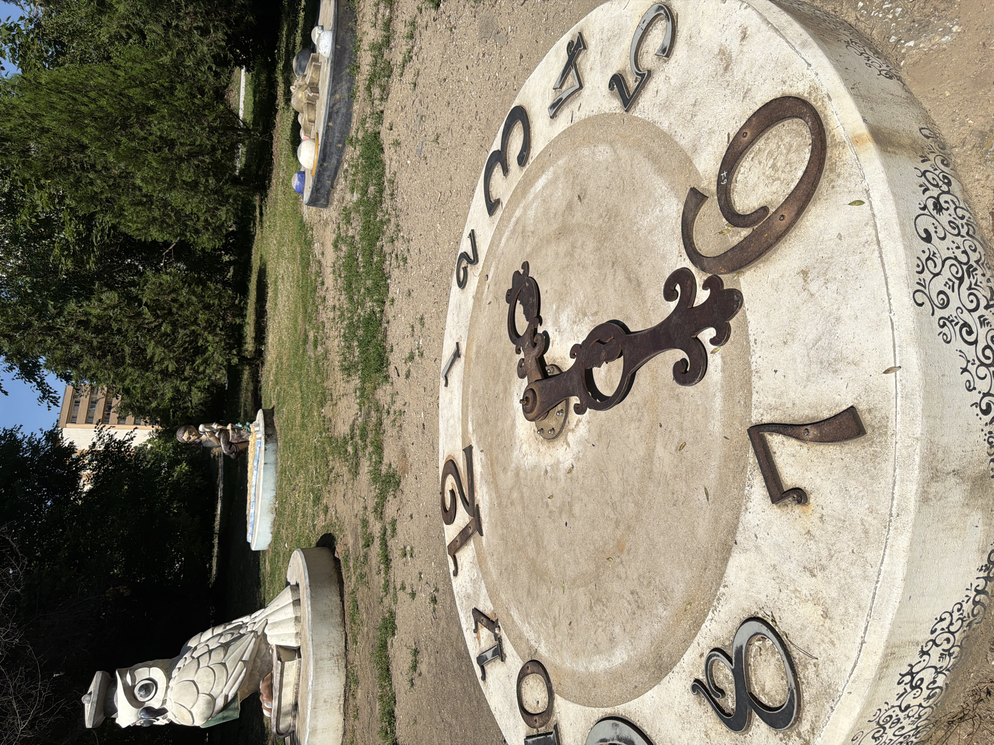The image depicts an outdoor area in Pazardzhik, Bulgaria, featuring a large, circular concrete structure designed to resemble a clock face. The clock has large, metal numbers indicating hours, with some numbers present and others missing or obscured. Decorative metal hands of a clock are attached to the center of the face. The clock structure is adorned with an ornate black scroll pattern along its base.
In the background, to the left of the clock, is a sculpture of a large, white owl wearing a graduation cap, perched on a book. Further in the background, a small sculpture of a boy sitting near a decorative fountain is visible. To the right, another decorative element with various objects, possibly stylized rocks or balls, is situated. The surrounding environment is a park-like setting with grassy areas, dirt paths, and mature trees. A multi-story building is visible in the distance, behind the trees. The scene is illuminated by natural sunlight.
