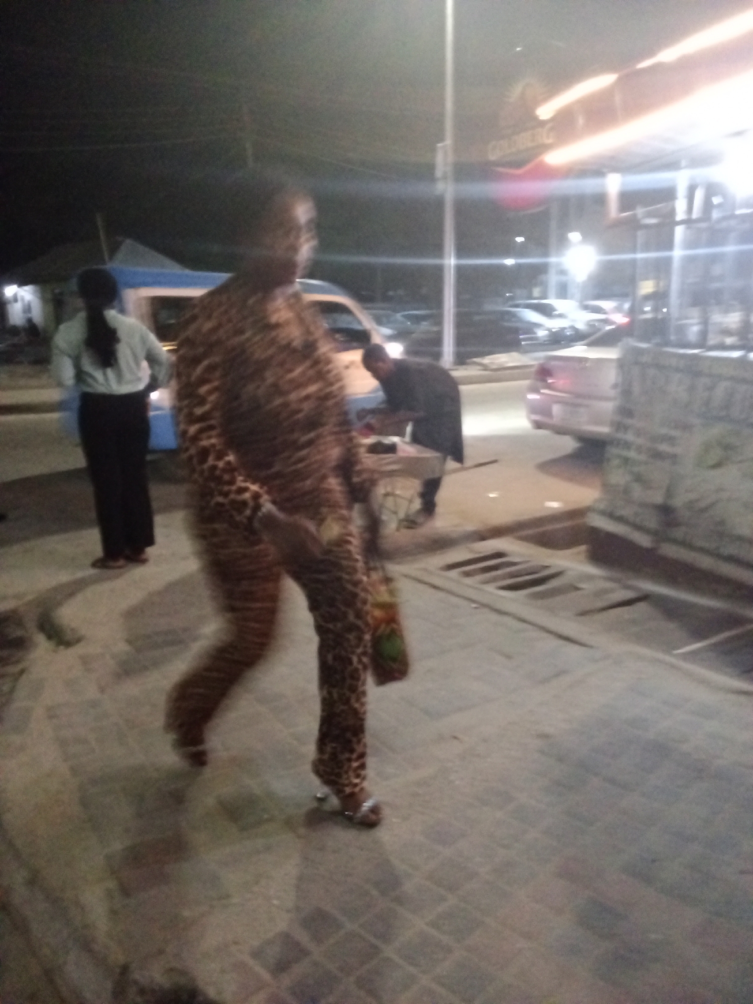 A street scene at night in Ikorodu, Nigeria shows a woman walking in the foreground wearing a brown and black patterned jumpsuit and silver sandals, carrying a bag. Behind her is a woman with long black hair, wearing a light colored top and dark pants. In the background, a man is bent over a small table. Several cars are parked along the street and a Goldberg beer sign is visible above a storefront. The area is paved with square stones, and there is a grated drain along the edge of the sidewalk.