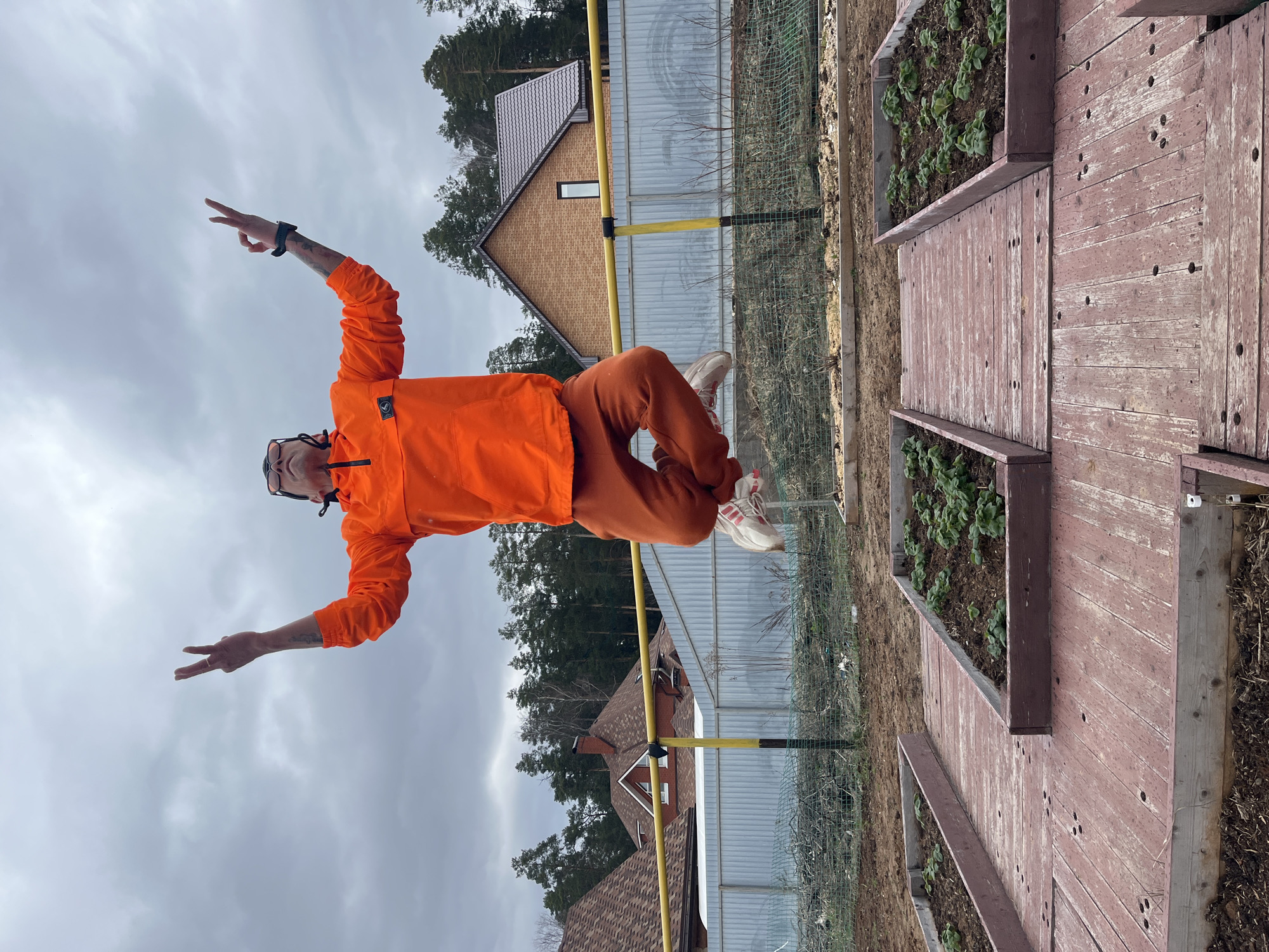 An adult individual is captured in a dynamic pose, balancing on a yellow horizontal bar outdoors in Chaikovsky, Russia. The person is wearing a bright orange hooded jacket and matching orange sweatpants, paired with white and red sneakers. They have glasses and visible tattoos on their forearms. Their left arm is raised with the hand forming a peace sign, while the right arm is also raised, with the index finger pointing upwards.
The background features an overcast, grey sky. Behind the balancing person, a sturdy yellow bar (possibly a gas pipe or part of a fence) runs horizontally across the frame. Further back, a metal fence and a mesh fence are visible. The landscape includes several residential houses with dark-tiled roofs, surrounded by tall evergreen trees and some bare deciduous trees, indicating a cooler season like early spring or late autumn. To the right, two elevated garden beds made of reddish-brown wooden planks are filled with soil and small green plants, likely young crops or ground cover. The ground surrounding the garden beds is dirt with some dried plant material. No discernible text is visible in the image.