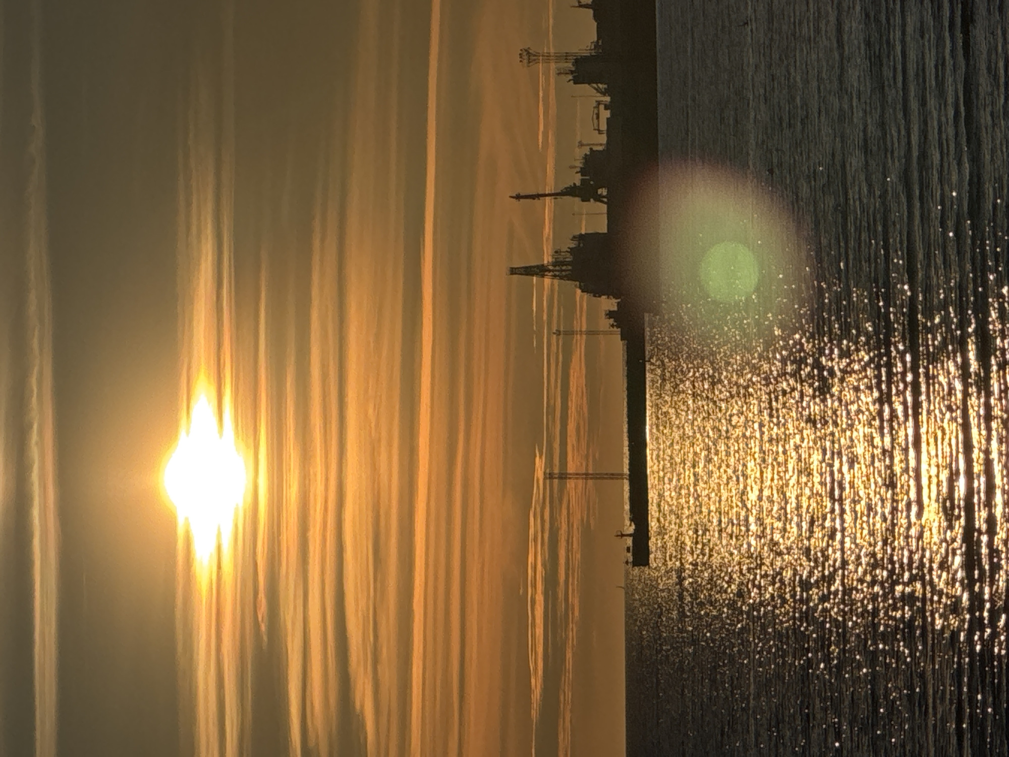 The image depicts a sunset over a harbor in Tallinn, Estonia. The sun, a brilliant white orb with golden rays, is positioned high in the sky, partially obscured by horizontal, orange-hued clouds. The water in the foreground is dark, reflecting the sunlight in a shimmering pattern of golden sparkles. Silhouetted against the bright sky are various port structures, including cranes and buildings, which form a distinct industrial skyline along the horizon. In the middle distance, a long, dark structure, possibly a pier or a docked ship, stretches across the water. A small boat is visible on the water to the left of this structure. The overall atmosphere is serene and industrial, illuminated by the warm glow of the setting sun.
