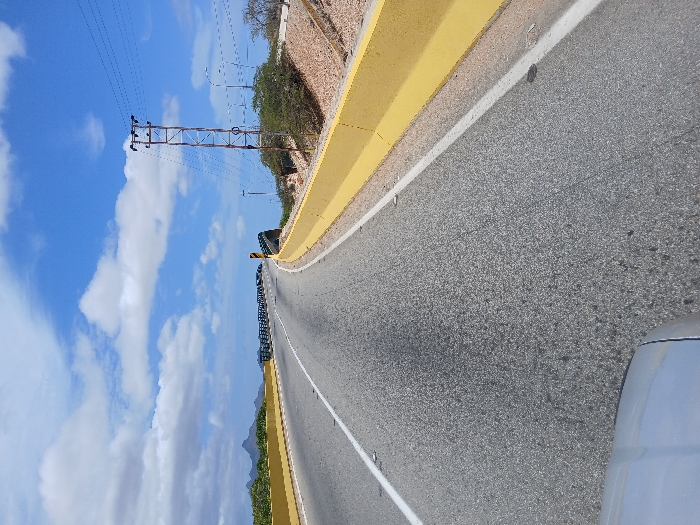 The image displays a paved two-lane asphalt road, likely a highway, located in the city of Santa María, Venezuela, under a blue sky with scattered white cumulus clouds during daylight hours. The road curves gradually to the right and appears to ascend, with bright yellow concrete barriers lining both sides. A solid white line delineates the left lane, and another solid white line marks the right edge of the road. On the left, the yellow barrier transitions into a dark-colored metal guardrail section, suggesting an elevated roadway structure. In the distant background on the left, low mountains or hills are visible. To the right of the road, a reddish-brown, dry embankment slopes upwards, covered with sparse green foliage. A prominent rusty metal lattice transmission tower stands on this embankment, supporting multiple overhead power lines. Additional streetlights on poles are visible further along the right side. The bottom center-left of the image shows the top edge of a light-colored vehicle's hood or dashboard, indicating the photograph was taken from inside a vehicle. No people, active vehicles, or specific actions are discernible within the frame.