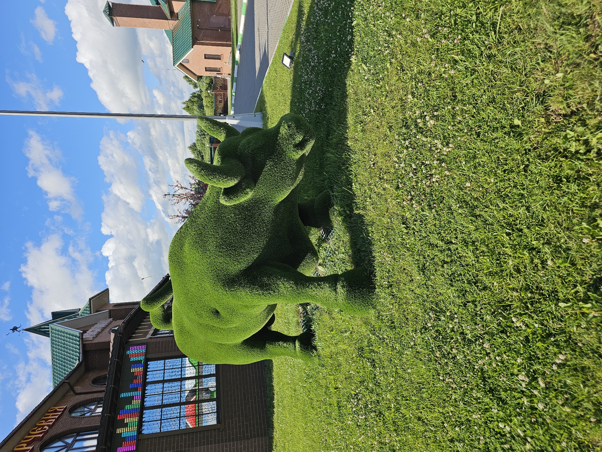 This image depicts an outdoor scene in the city of Ilyinskoye, Russia, on a sunny day with a partly cloudy blue sky. In the foreground, a large, artificial bull sculpture covered in green artificial grass stands on a lush green lawn dotted with small white flowers. The bull is realistically sculpted, with visible legs, body, head, and horns. To the left of the bull, a brick building with arched windows and a green tiled roof is visible. One window has a colorful LED display resembling an equalizer. To the right, a paved pathway borders the lawn, and further back, another brick building with a green roof and a chimney is present, along with some trees and a fence. A tall, slender pole stands near the bull sculpture, and a spotlight is visible on the pathway.