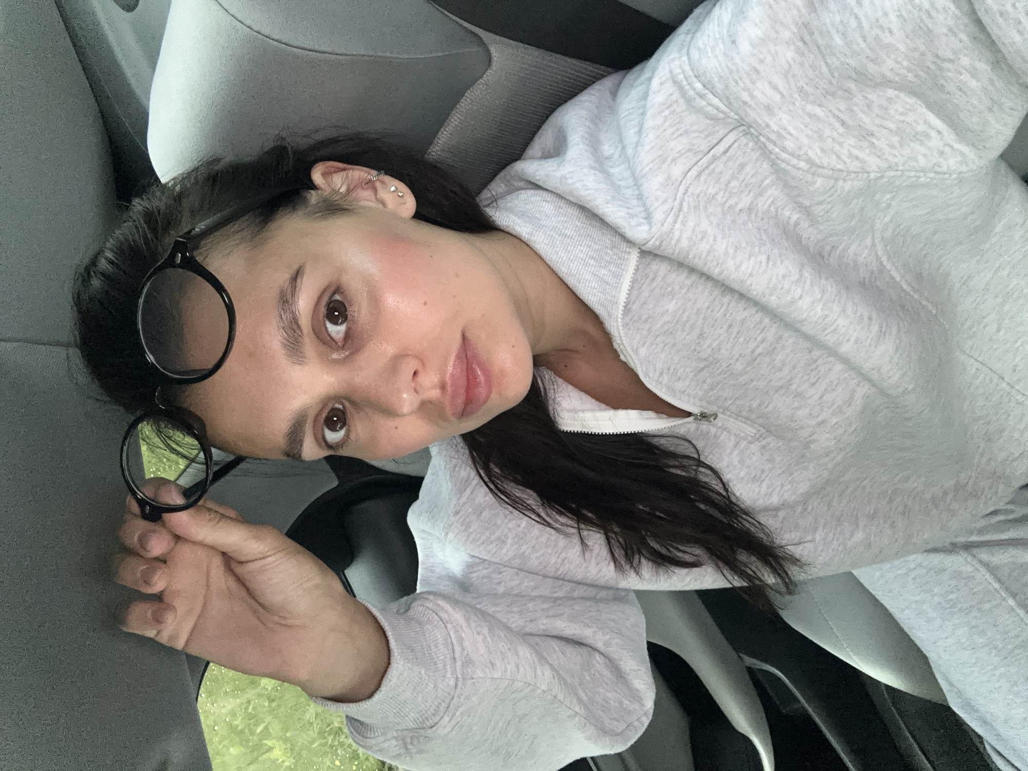 A woman with dark hair is sitting in the back seat of a car, looking at the camera. She is holding a pair of round-framed glasses above her head with her right hand. She is wearing a light grey zippered sweatshirt. The car's interior is visible, including a grey headrest and seat. Outside the car window, green foliage is visible. The image appears to be a selfie taken from inside the vehicle. There is no information in the image to suggest the location is the city of Perm, Russia.