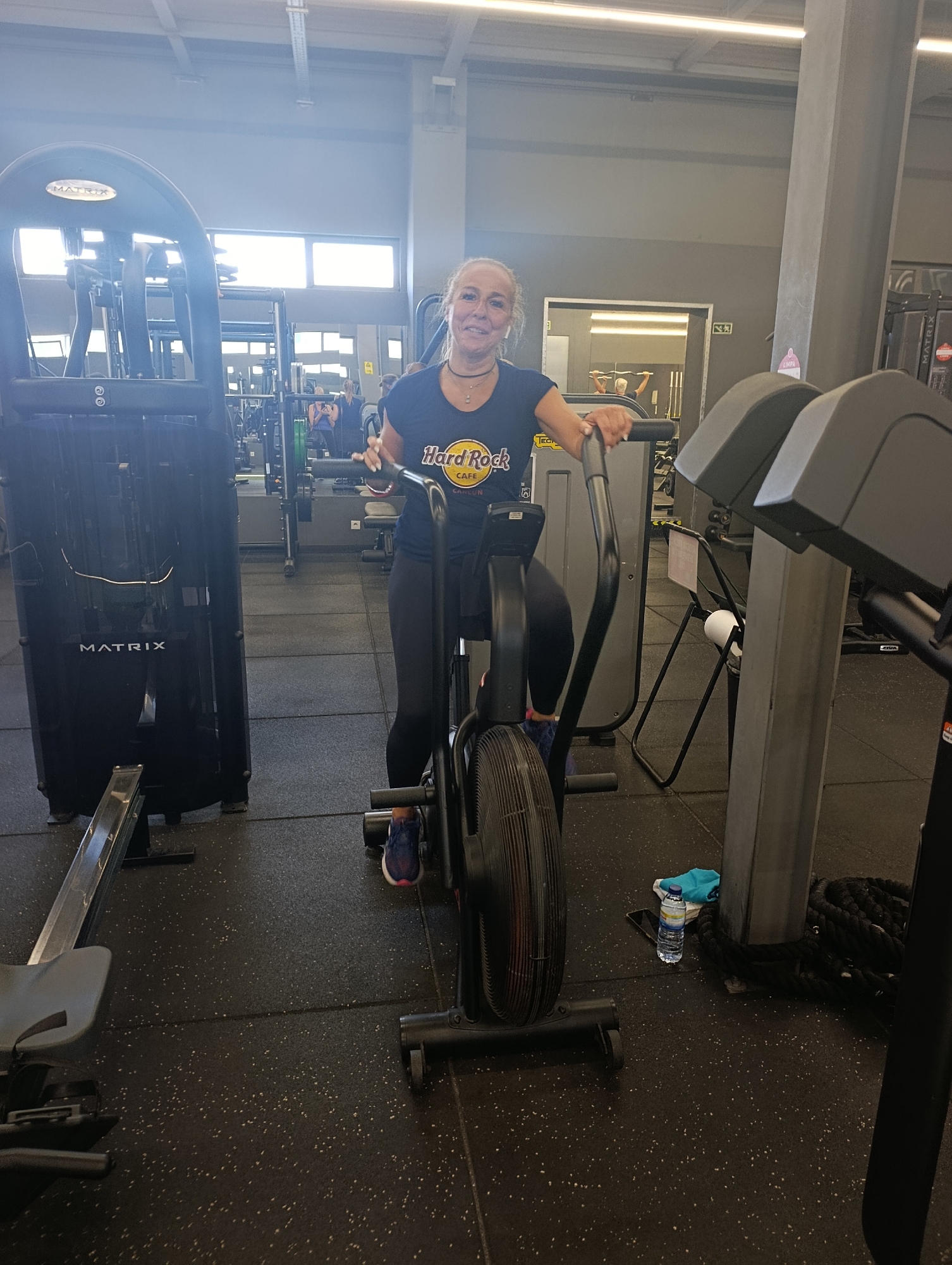 In Setúbal, Portugal, a woman is pictured in a gym, riding an air bike. She is wearing a dark blue t-shirt with a "Hard Rock Cafe" logo and black leggings. She has blonde hair tied back and is wearing a choker necklace. Her hands are on the handlebars of the air bike. The gym has a grey floor with speckles and various exercise equipment visible, including a rowing machine to the left, an air bike in the foreground, and other weight machines in the background. There are windows with natural light coming in from the top and the back wall appears to be a dark grey. A water bottle and a blue towel are on the floor near the air bike.