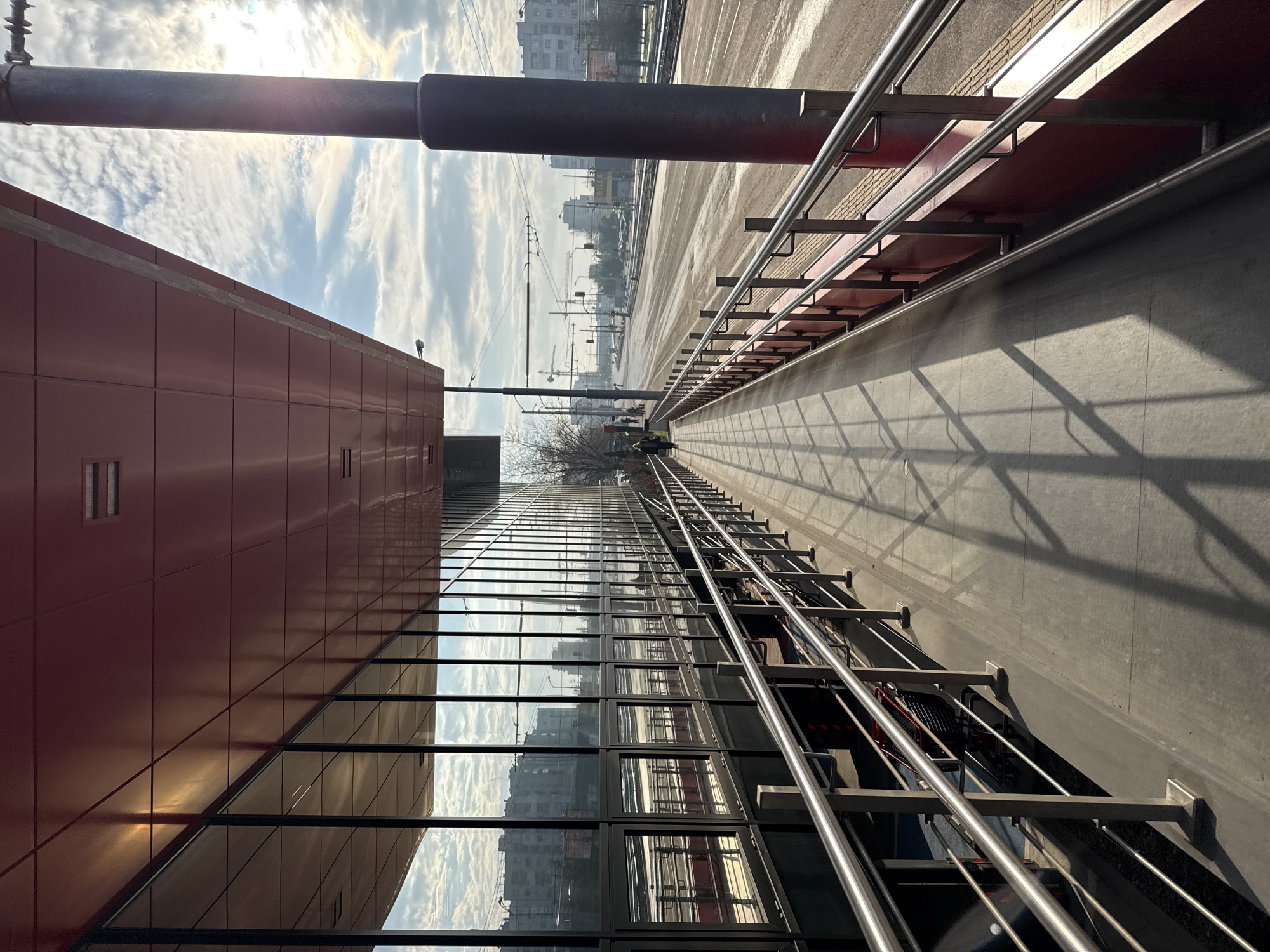 The image shows a pedestrian walkway alongside a building in what appears to be an urban setting. The building has a red paneled section and a large glass facade reflecting the sky and surrounding buildings. A metal railing runs along the walkway. In the distance, other buildings can be seen in Sofia, Bulgaria, with an overhead tram wire and utility poles. The walkway itself is paved with shadows from the overhead structure.
