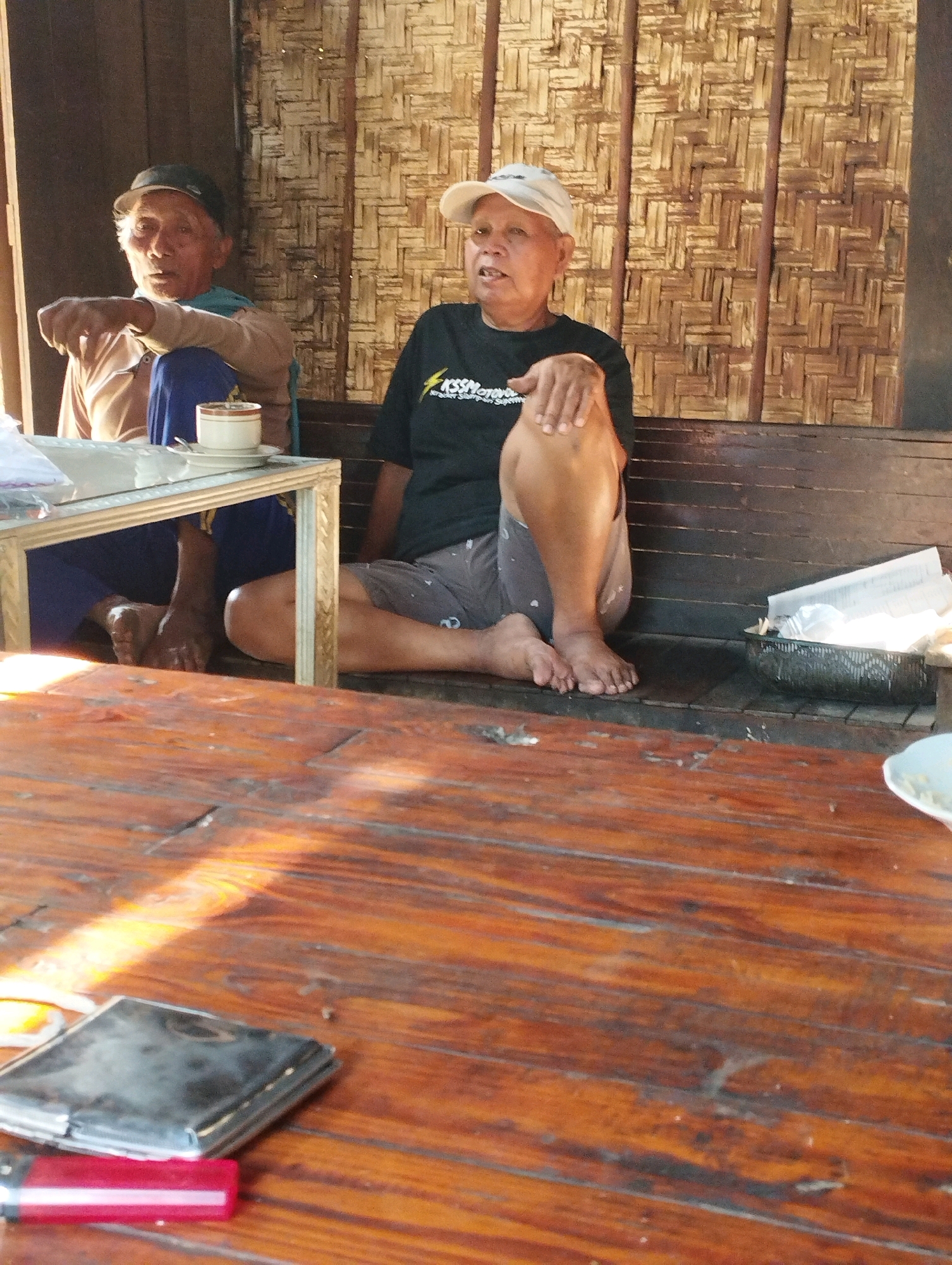 Two elderly men are seated indoors. The man on the left wears a light brown shirt and a dark baseball cap. He has a blue garment draped over his shoulders. He is leaning on a small table with a white cup and saucer. The man on the right wears a white baseball cap and a black t-shirt with white text. He is wearing grey shorts. They are sitting on a wooden bench against a wall made of woven bamboo. A black plastic basket containing white paper is to the right of the man on the right. In the foreground, a wooden table with a visible grain runs across the image. On the table are a metallic case and a red lighter. The lighting suggests the scene is illuminated by natural light coming from the left. The location is the village of Wedarijaksa, in the Pati Regency of Central Java, Indonesia.