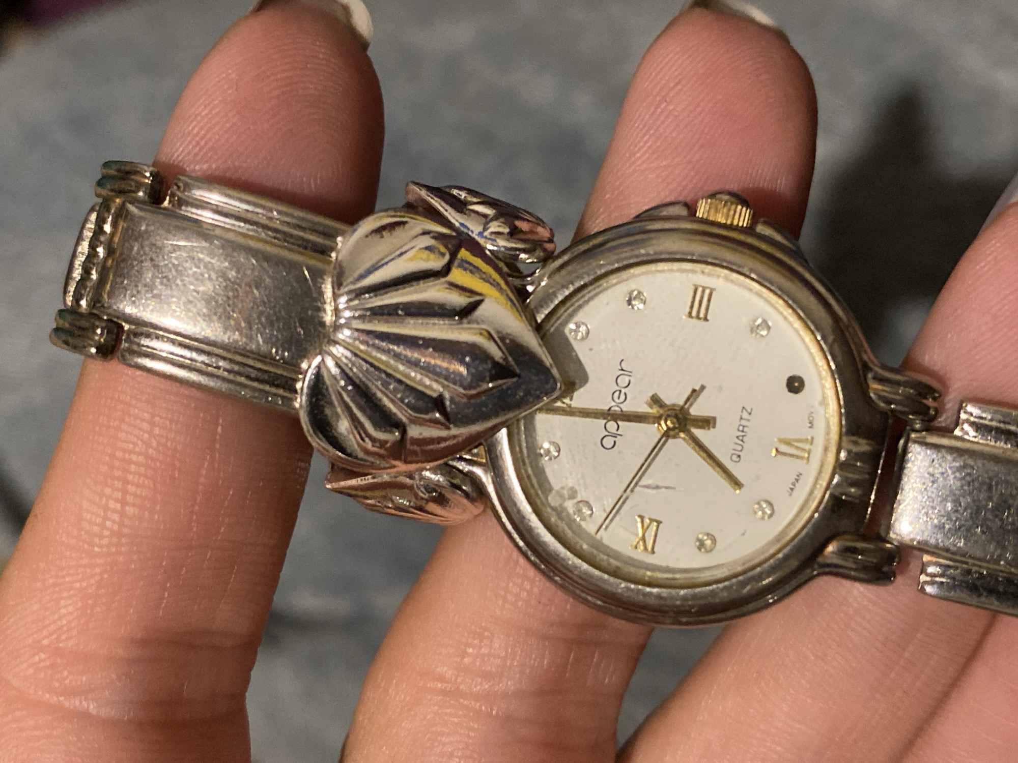 A person is holding a silver-colored watch with a white face. The watch features Roman numerals and the brand name "Appear Quartz" on the dial. The band is made of silver-colored metal and includes a decorative, shell-shaped element. The watch appears to be worn. The background is a blurred, neutral tone. The location in Monte Grande, Argentina, cannot be determined from the image.