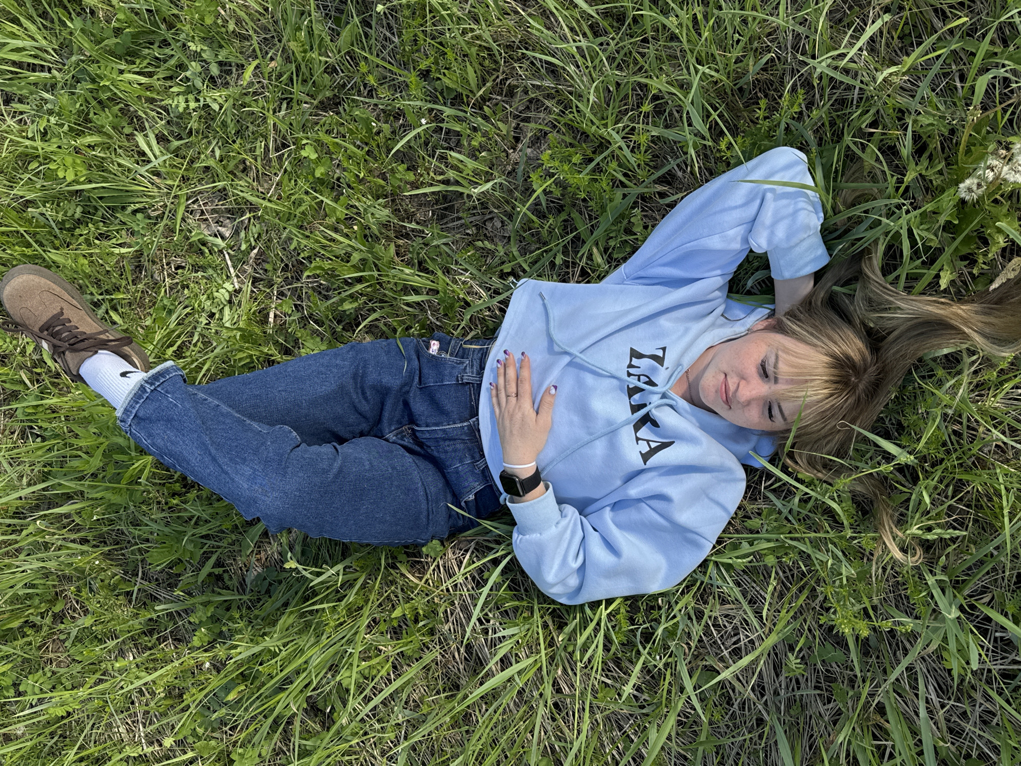 A young person is pictured lying on their back in vibrant green grass, looking up towards the camera with a subtle smile. The scene is an outdoor setting, likely a field or meadow, with varying lengths of grass and some dried patches of earth visible. The person is wearing a light blue hooded sweatshirt with dark lettering on the front, which appears to spell "ZARA" in part. They are also dressed in blue denim jeans and brown lace-up shoes with white socks that have a small, dark logo near the ankle.
Their left arm is bent, resting across their stomach, revealing a watch with a black band on their wrist and a ring on their finger. Their nails are painted a light purple. Their right arm is bent with the hand near their head, and their light brown hair is spread out on the grass around their head. The lighting suggests it is daytime, likely sunny, given the clear visibility and brightness of the scene. In the upper right corner of the image, some white, fluffy seed heads, possibly dandelions, are visible among the grass. The overall impression is one of relaxation and calm in a natural environment. The location context provided is Markovskiy, Russia.
