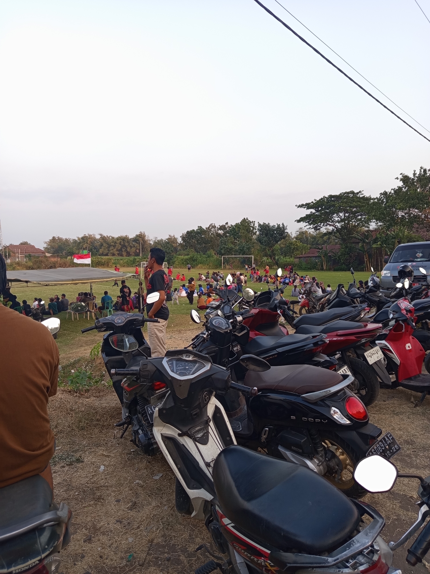 The image depicts an outdoor scene with a gathering of people on a grassy field, likely a sports field, in Mrican, Indonesia. In the foreground, several motorcycles are parked. The sky is overcast with a pale, hazy quality. In the background, a soccer goal is visible, and some individuals are engaged in activities on the field. A group of people are seated on the grass, some are standing, and a few are gathered around tables. A red and white flag, possibly the Indonesian flag, is flown from a pole. Trees and buildings can be seen in the distance. The overall atmosphere suggests a communal event or a casual gathering.