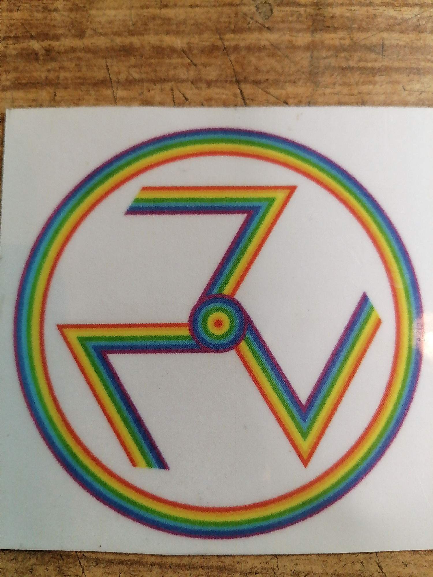 The image shows a sticker with a rainbow-colored symbol resembling a swastika on a white background. The symbol is composed of three angular arms that radiate from a central circle, also filled with rainbow colors. Each arm is outlined and filled with concentric rainbow stripes. The outer edge of the sticker is a complete rainbow circle. The sticker is placed on a wooden surface with a visible grain. The image is taken from a slightly elevated angle, looking down at the sticker. There are no people or any other objects in the image. There is no information in the image that would allow for the determination of its location in Rostov-on-Don, Russia.
