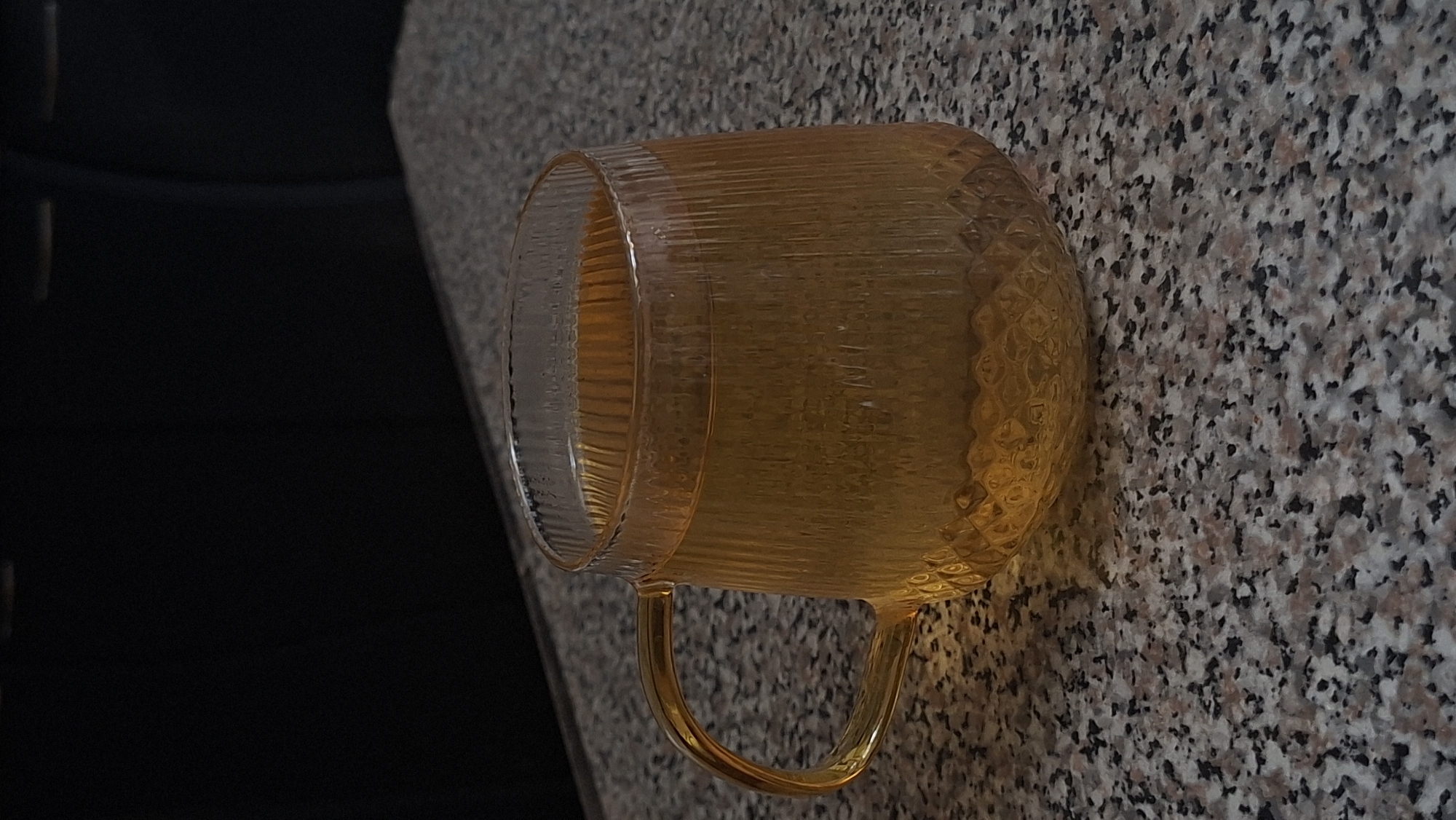 A clear glass mug filled with a golden-yellow liquid, likely tea, sits on a speckled granite countertop. The mug has a ribbed texture and a curved handle. The background is dark and out of focus, suggesting an indoor setting. The image does not contain any information that would allow for determination of its location in the city of Myadzyel, Belarus.