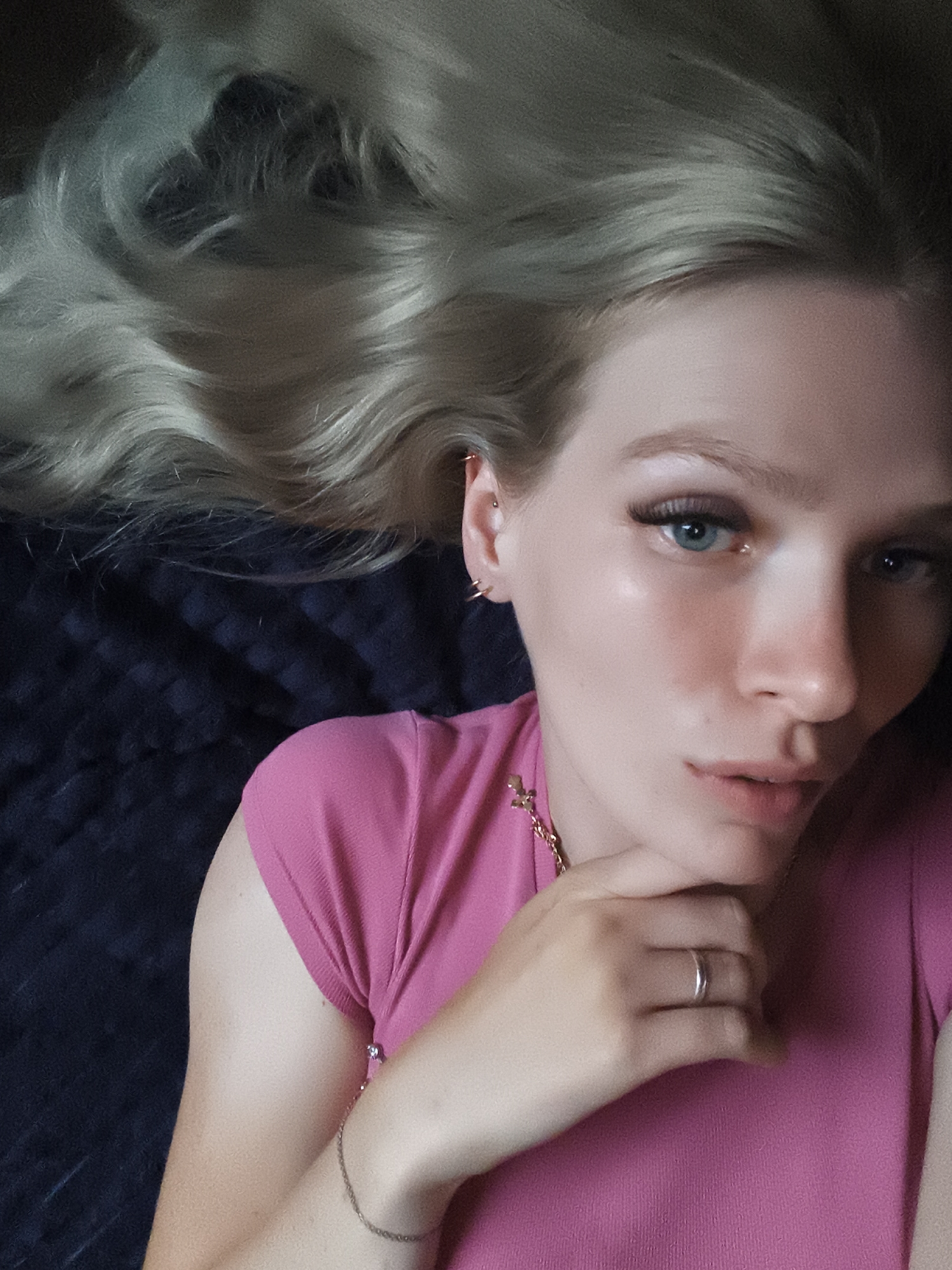 The image depicts a close-up, overhead shot of a young woman lying down, with her blonde hair spread across a dark, textured surface, possibly a bed or couch. She is looking directly at the camera with a neutral expression. The woman is wearing a bright pink, short-sleeved top. A gold chain necklace with a cross pendant is visible around her neck. Her right hand is resting on her chin, and she is wearing a silver ring on her left ring finger. A thin bracelet is visible on her left wrist. The lighting suggests an indoor setting, possibly natural light, with a slight sheen on her skin. The overall impression is of a casual, self-portrait. There is no discernible location in Русский Вожой, Russia.