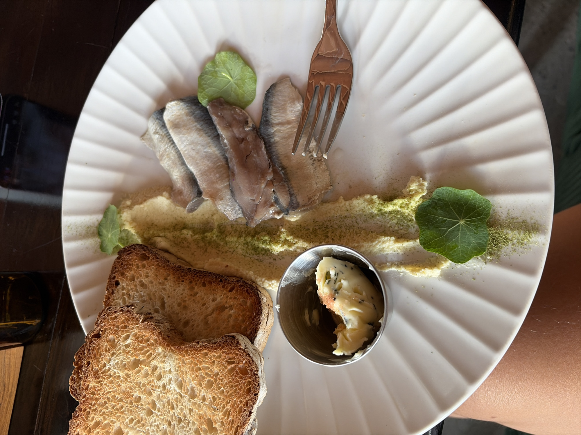 An overhead, close-up shot shows a white, fluted plate of food. Two slices of golden-brown, toasted bread are positioned in the upper left quadrant of the plate. Adjacent to the toast, in the lower left, is a small, silver metal ramekin filled with a pale yellow spread, possibly butter, flecked with dark specks.
A smear of pale yellow purée, dusted with green powder, runs horizontally across the plate, separating the toast and butter from the fish. In the upper right quadrant, arranged in a slight curve, are four fillets of cured herring, each with a silver skin and pale pink flesh. A small, green leaf rests on the purée to the right of the herring. Another similar leaf is situated below the herring on the left. A silver fork with three tines rests on the plate, pointing towards the herring.
The plate is set on a dark wooden table. A partially visible brown glass bottle is present in the background, and the reflection of a cell phone screen is visible on the table's surface. The lower portion of the image shows the skin of a person's arm and thigh.
The scene is set in a restaurant in North Kuta, Indonesia.