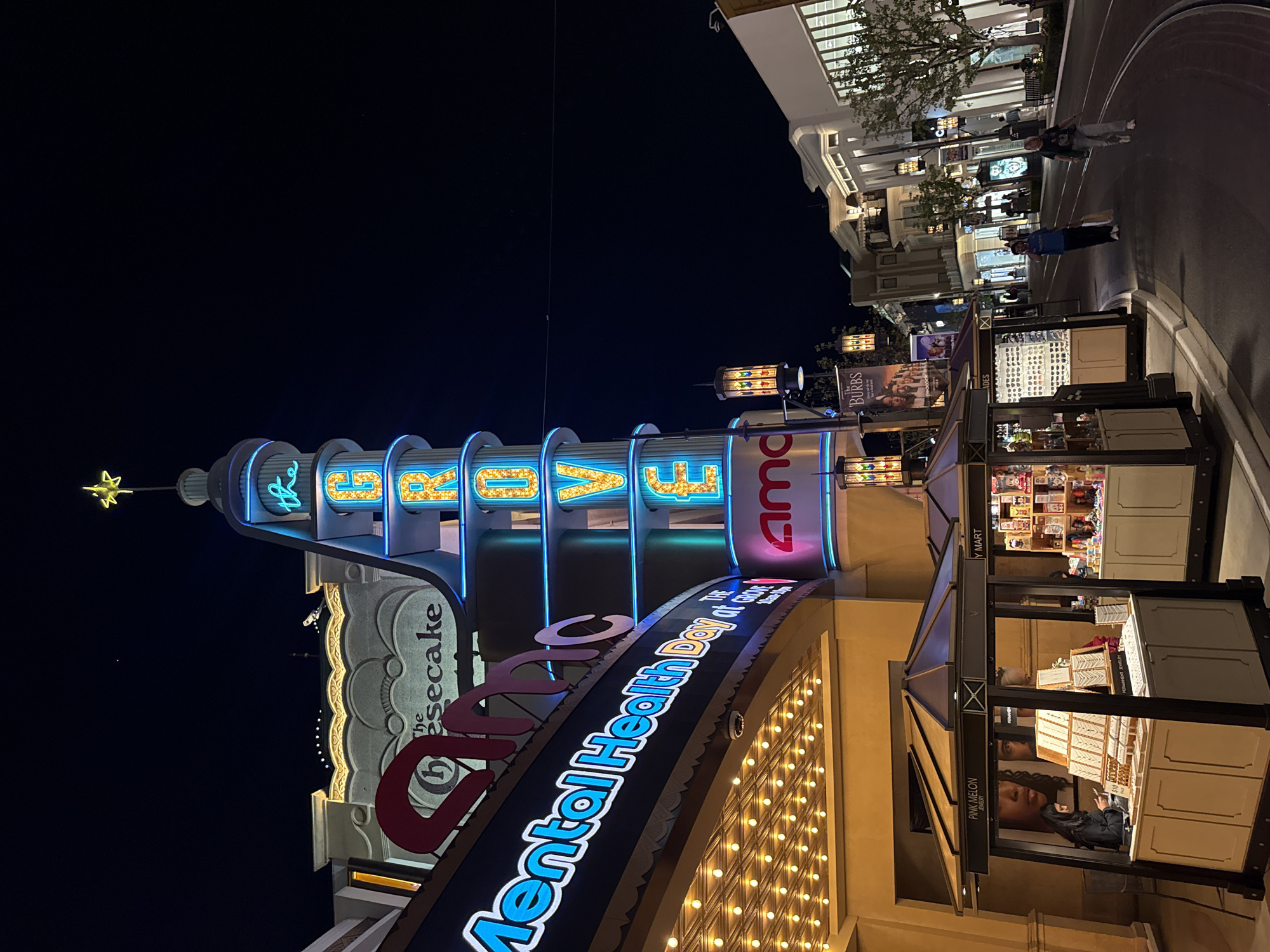 This nighttime shot captures a bustling scene at The Grove in Los Angeles, California. The iconic "The Grove" sign is prominently displayed atop a stylized tower. To the right of the sign is an AMC movie theater sign. A large sign below promotes "Mental Health Day at Grove." Buildings and storefronts line the background, illuminated with various lights and signage. Small retail kiosks are visible, with vendors. People can be seen walking on the pavement, suggesting activity and commerce. The sky is dark.