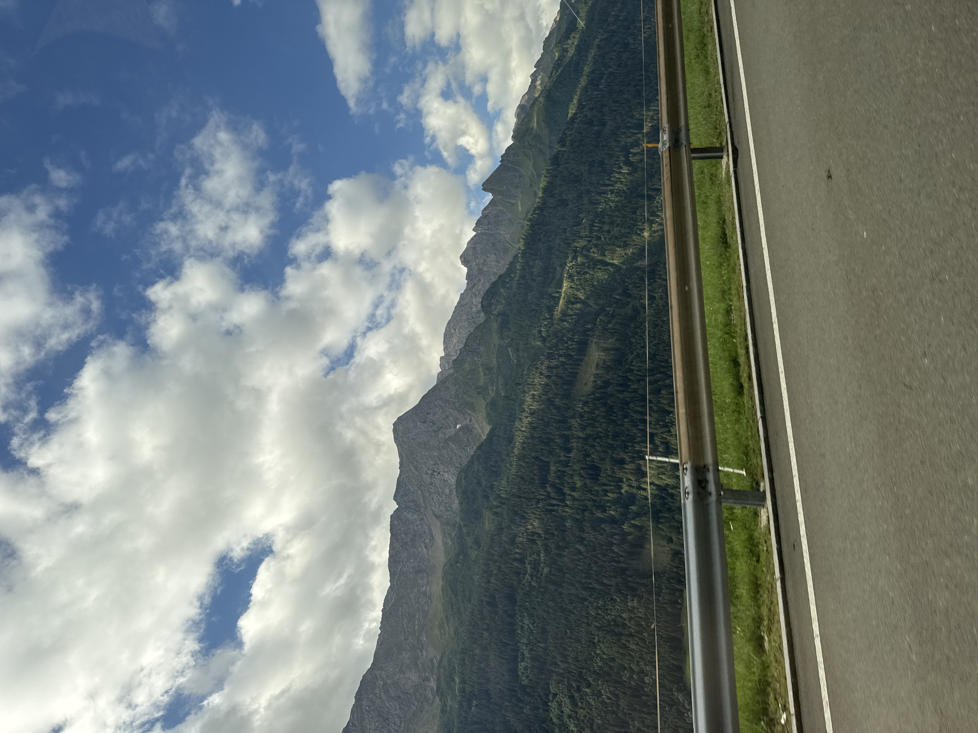 The image captures a scenic view of a mountainous landscape in Airolo, Switzerland, as seen from a roadside. In the foreground, a portion of an asphalt road with a white lane marker is visible. A silver guardrail with visible bolts runs parallel to the road, separating it from a grassy embankment. Beyond the embankment, a dense forest of evergreen trees covers the lower slopes of a rugged mountain range. The peaks of the mountains are rocky and appear to be at a higher elevation, with some patches of snow or lighter colored rock. The sky above is a vibrant blue, dotted with fluffy white clouds, suggesting clear weather. The overall impression is one of natural beauty and a sense of travel along a scenic route.
