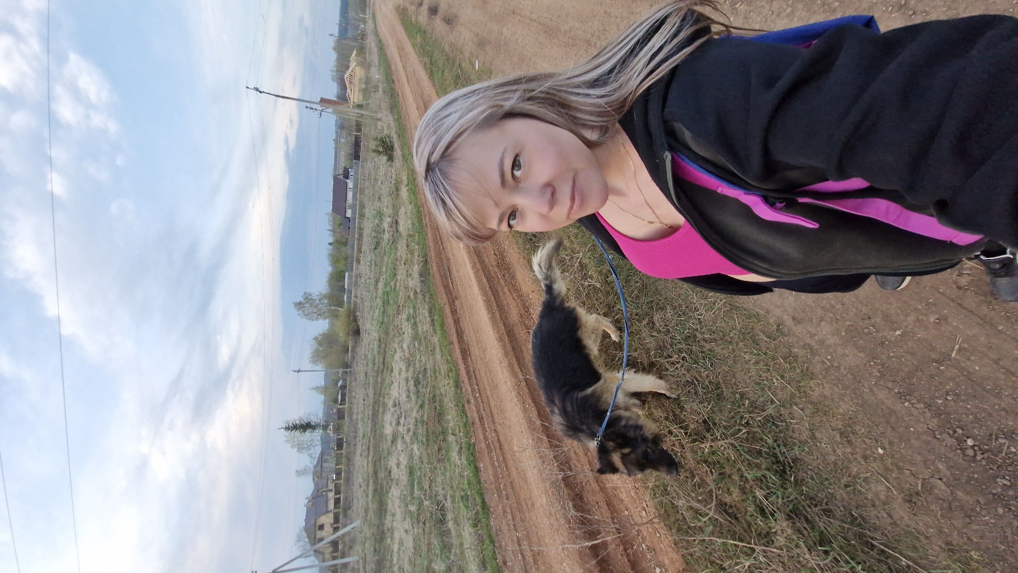 A woman and a dog are visible on a dirt road near Otchor, Russia. The woman is fair-skinned with blonde hair and is wearing a pink tank top and a black hoodie with pink accents. She is holding a leash attached to a medium-sized dog with dark and light brown fur. The road has tracks and vegetation along the sides. Houses and utility poles are visible in the distance. The sky is partly cloudy.