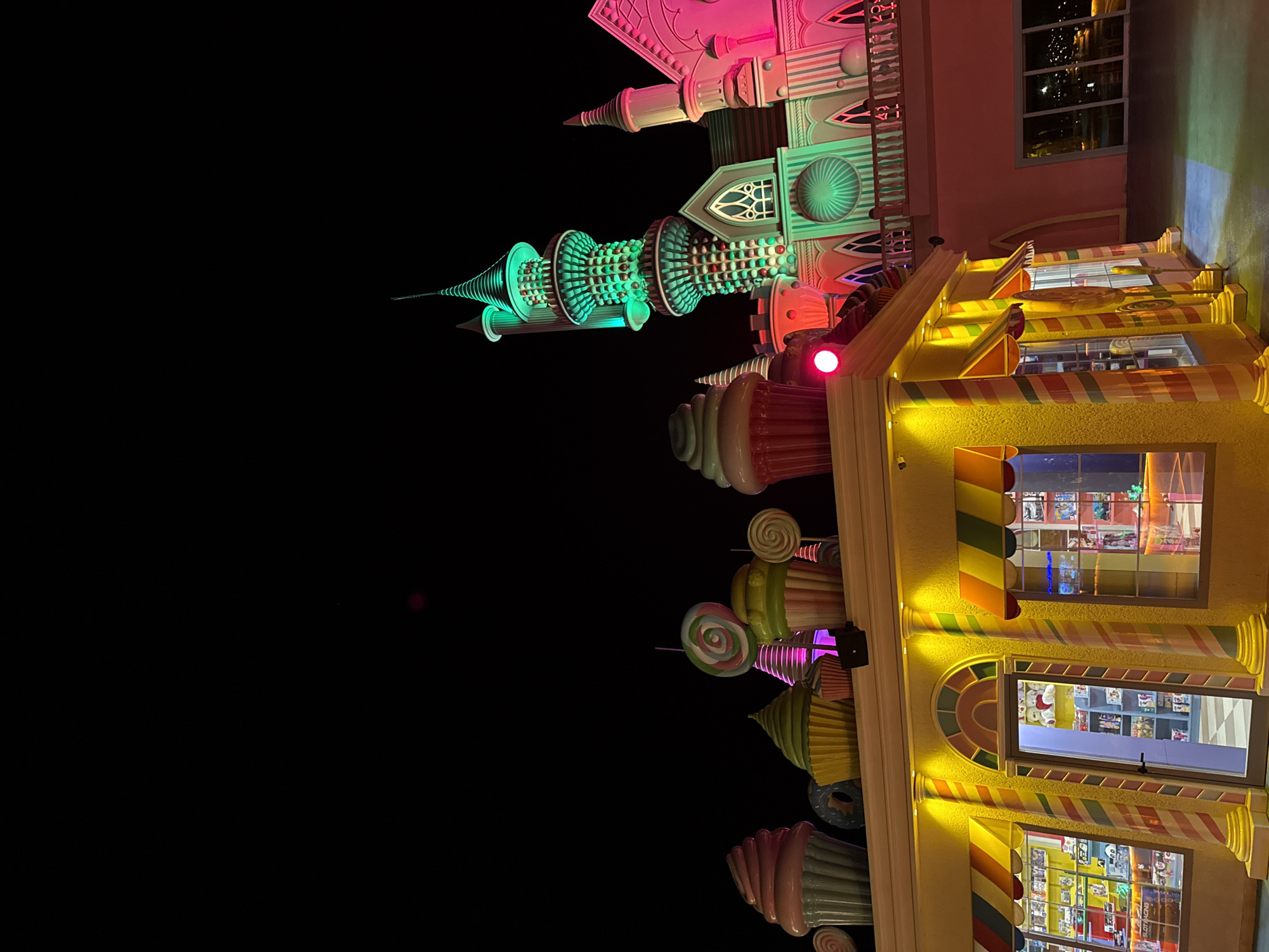 The image shows a whimsical candy-themed attraction at night, likely part of The Land of Legends theme park in Bodrum, Turkey. A brightly lit, yellow candy shop with colorful awnings and striped pillars is in the foreground. Behind it, a fantasy castle-like structure is illuminated with vibrant pink and green lights. Various oversized candy decorations, including cupcakes and lollipops, are visible along the rooftop of the shop and the castle. The dark sky suggests it is nighttime.