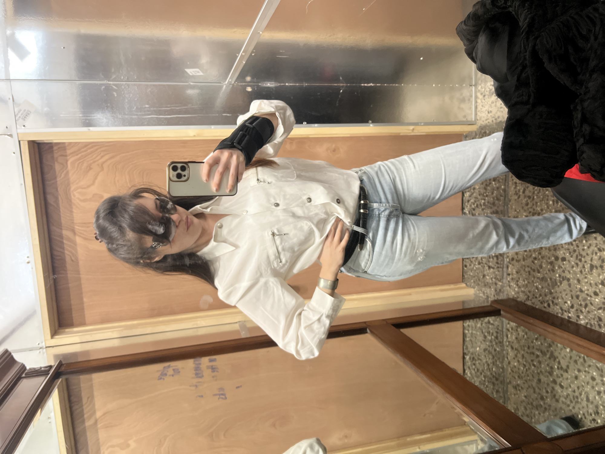 The image depicts an interior scene, likely a fitting room or utilitarian space within a commercial establishment in New York City, United States of America.
A single female individual is centrally positioned, taking a mirror selfie. She has long, dark hair, partially pulled back, with bangs, and wears dark sunglasses with metallic accents on the temples. Her attire consists of a white long-sleeved collared shirt with two breast pockets, one featuring a zipper, and light-colored buttons. She wears light blue, distressed denim jeans and a black belt with a silver buckle and multiple small studs. A silver-colored cuff bracelet is visible on her left wrist. A black wrist brace is worn on her right wrist, beneath the shirt cuff, as she holds a light-colored smartphone with a triple-lens camera in her right hand to capture the image. Her left hand rests on her left hip.
The background includes a light brown or wooden-paneled wall or door on the left, reflecting faint, illegible mirrored text. To the right, a metallic wall, possibly aluminum or galvanized steel, shows vertical seams and minor surface streaks. A dark-wood framed full-length mirror stands on the left, reflecting parts of the room. The floor is a speckled terrazzo or concrete surface. In the lower right corner, a pile of dark-colored fabric, possibly clothing, with a small visible section of red material, rests on an unseen object.