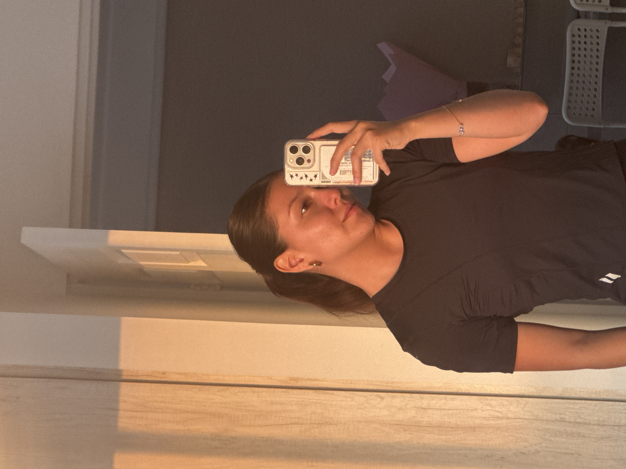 A young woman is captured in a mirror selfie, holding a smartphone to photograph her reflection. The image is taken indoors, likely during golden hour, as warm, directional light bathes parts of the wall and floor in an orange glow.
The woman has dark hair pulled back, revealing a small earring in her visible ear. She wears a plain black short-sleeved t-shirt with a double-stripe logo on the right sleeve. A delicate bracelet with three small, round beads adorns her right wrist. Her expression is neutral to slightly pensive as she looks towards the phone.
She holds a silver or light-colored smartphone with a clear case. The back of the case features a white rectangular insert or design. The phone's camera module, visible in the upper left of the case, has three lenses and a flash. Below the camera, several small star-shaped symbols are visible on the white insert. Some text is printed on the white part of the case, but it is largely illegible due to reflections and image quality, though "qalbu.w" is vaguely discernible among other obscured characters.
The setting appears to be a room with light-colored walls and a reflective floor, possibly wood or tile, catching the warm light. In the background, beyond the woman, parts of a dark wall or surface are visible, along with a grid-patterned object, possibly a storage unit or chair, and a soft purple object. The lighting creates distinct shadows and highlights throughout the scene. The context provided implies the photo was taken in San Francisco Ocotlán (Ocotlán), Mexico.