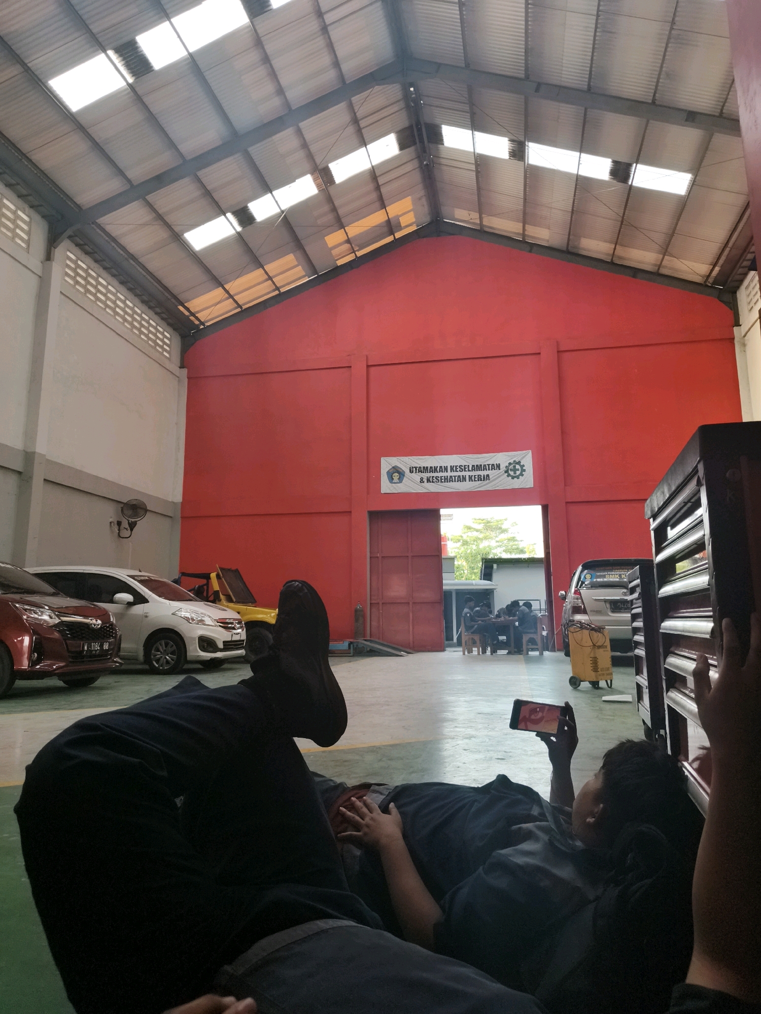 The image depicts the interior of a workshop, possibly a car repair or vocational school, in Katrungan, Indonesia. The workshop has a high, corrugated metal roof with several translucent panels allowing natural light to enter. The walls are painted in sections of light gray and a vibrant red. A banner displaying the text "UTAMAKAN KESELAMATAN & KESEHATAN KERJA" (Prioritize Safety & Health at Work) with accompanying icons is visible on the red wall.
Several vehicles are present, including a red car in the foreground, a white car, and a yellow open-top vehicle. Further in the background, another silver car is visible near a red door. A yellow cart with equipment is positioned near the silver car. A group of individuals appear to be gathered around a table inside the workshop through an open doorway.
In the foreground, two individuals are lying down on the floor. One person, whose legs and feet are prominently in view, is wearing dark pants and appears to be resting. The other person, facing away from the camera, is looking at a smartphone held in their hand. Their posture suggests they are also relaxing. The floor is a combination of green and gray concrete. The overall impression is a casual moment within a working environment.