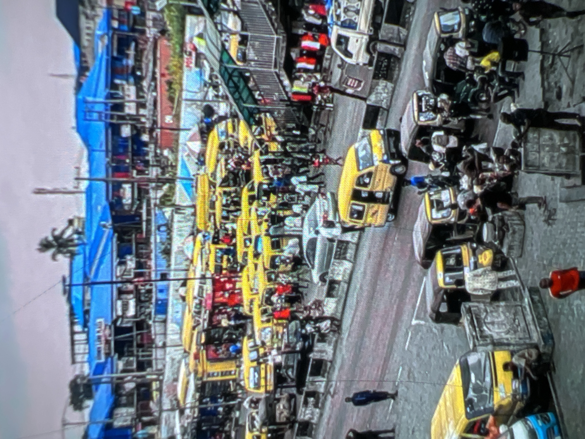 The image depicts a busy street scene in Lagos, Nigeria, likely a market area or transportation hub. The foreground shows a variety of vehicles, including yellow minibuses (danfos), a white truck, and several three-wheeled auto-rickshaws (keke napep). Numerous people are visible, some standing, some walking, and others gathered around the vehicles. Many appear to be passengers or vendors. The middle ground is dominated by more danfos and pedestrians. In the background, a building with a bright blue roof stands out, possibly a market or commercial complex. A palm tree is visible to the left of the building. The overall impression is one of a bustling, dynamic urban environment characteristic of Lagos.