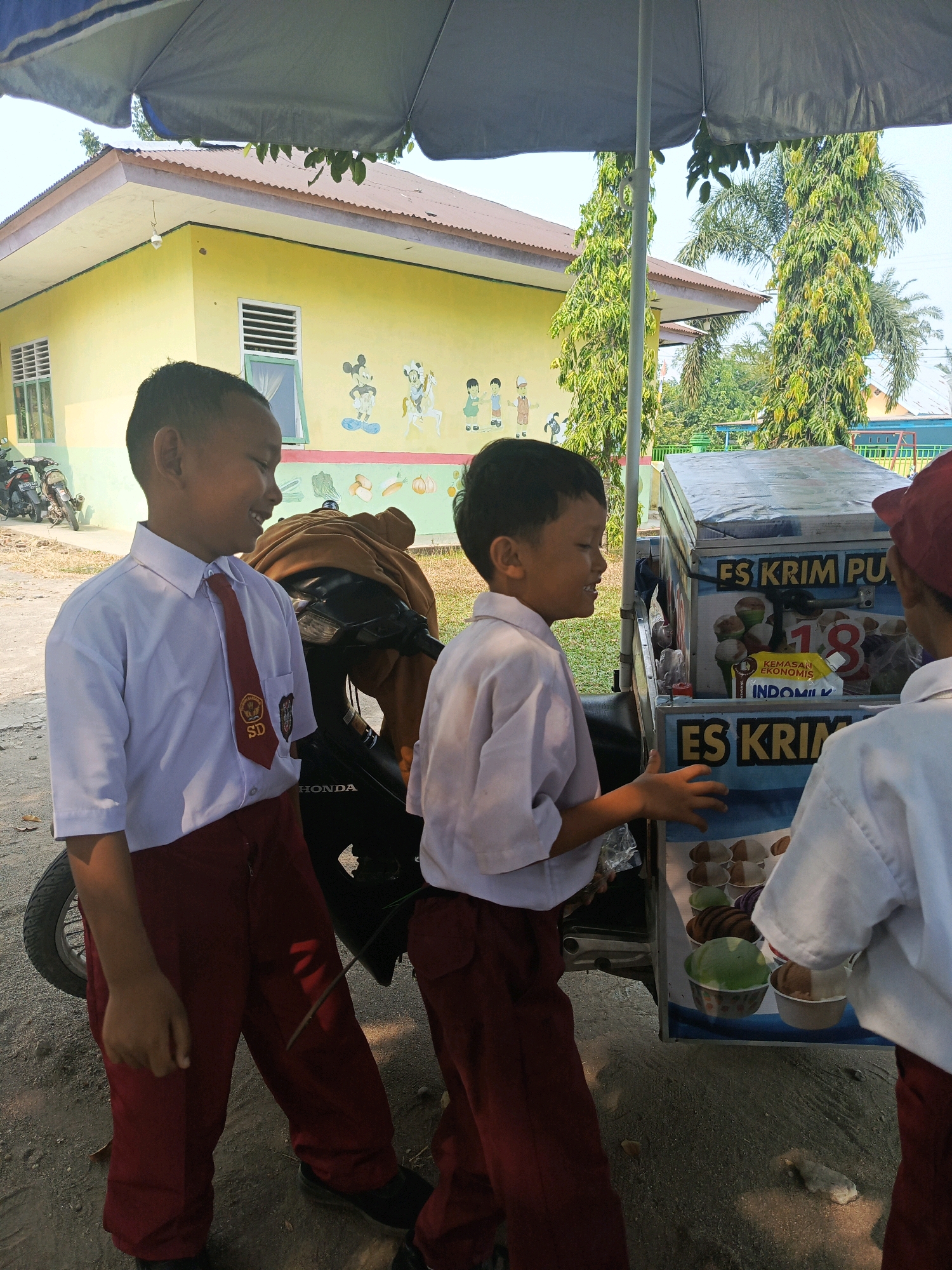 In Karya Dadi, Indonesia, three young boys in school uniforms stand near an ice cream cart under a blue umbrella. Two boys, wearing white shirts and red pants, are on the left. The boy on the far left smiles broadly, his red tie with a school emblem visible. Another boy, positioned between the first and the cart, faces the cart and gestures with his hands. The third boy, on the right, also in a white shirt and red pants, wears a red cap and faces the cart. The ice cream cart has "ES KRIM PU" and "ES KRIM" written on it, with various ice cream cups visible inside. Behind them, a bright yellow building with colorful cartoon murals of Mickey Mouse and other characters is visible, along with trees and a motorcycle. The ground is dirt and gravel.