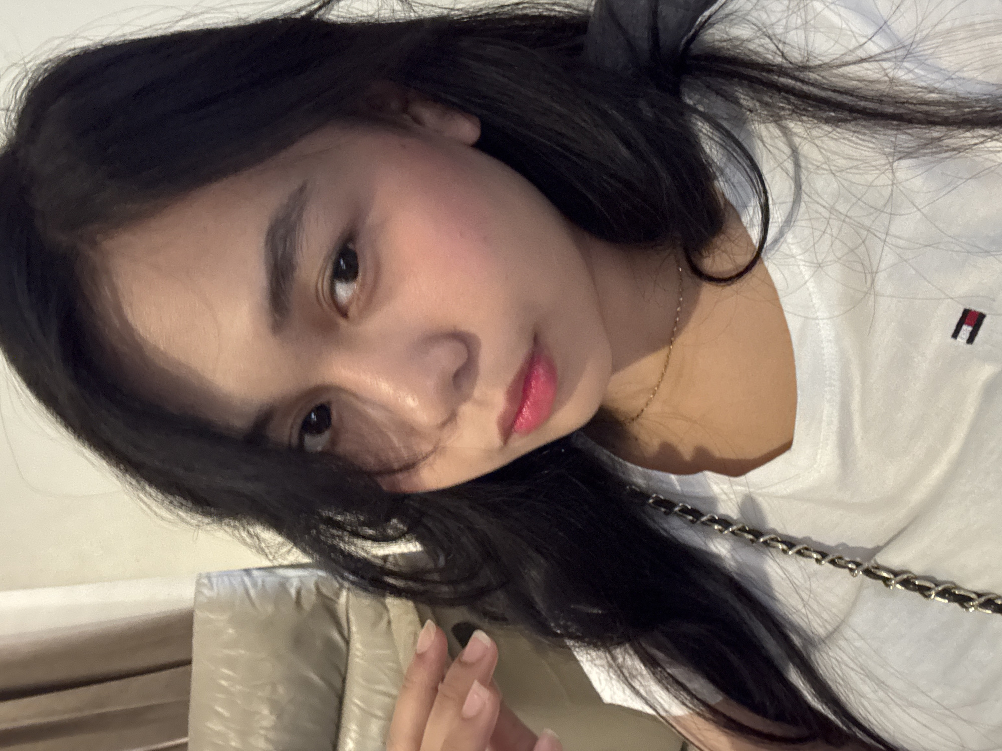 A young woman with long, dark hair and fair skin is pictured in a close-up selfie. She is wearing a white t-shirt with a Tommy Hilfiger logo on it and a delicate gold necklace. Her makeup includes pink blush and vibrant pink lipstick. She is looking directly at the camera with her right eye slightly more visible than her left due to her head being turned. Her left hand is positioned near her face, with her fingers slightly curled. In the background, a portion of a beige leather couch is visible, along with a beige curtain or wall. The lighting suggests an indoor setting. The image does not provide any explicit information about its location within Davao City, Philippines.