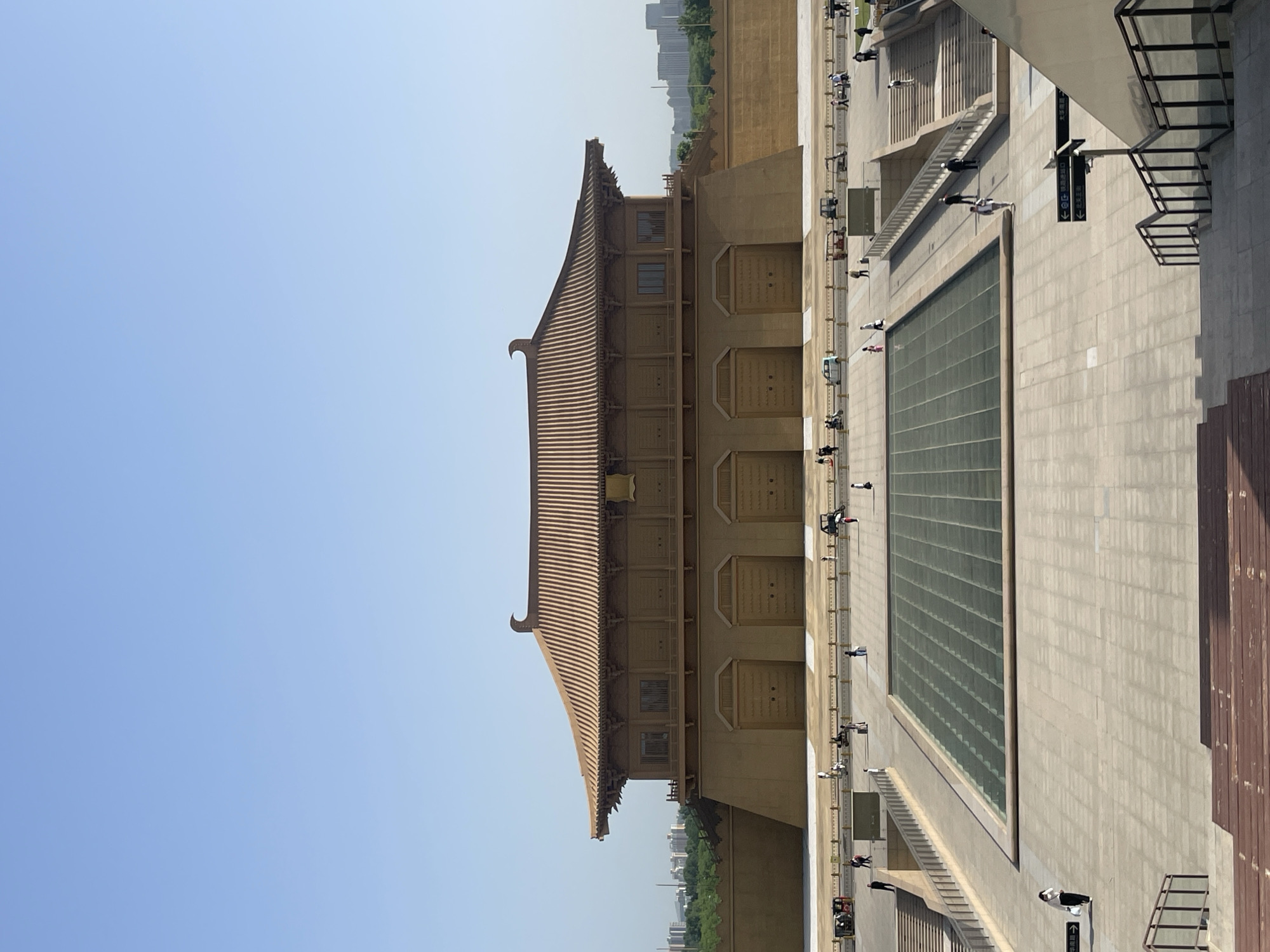 The image captures a view of the Yongning Gate, also known as the South Gate, in the city of Xincheng District, China. The architecture of the gate is a classical, multi-tiered structure with a tan hue and a traditional tiled roof. Clear blue sky dominates the upper part of the frame. At the base of the gate, there is a public square with a large glass panel and a concrete floor. People are walking through the square. Stairs and railings are visible alongside the square. Modern buildings are visible in the background, contrasting with the traditional architectural style of the gate. The lighting suggests a sunny day.