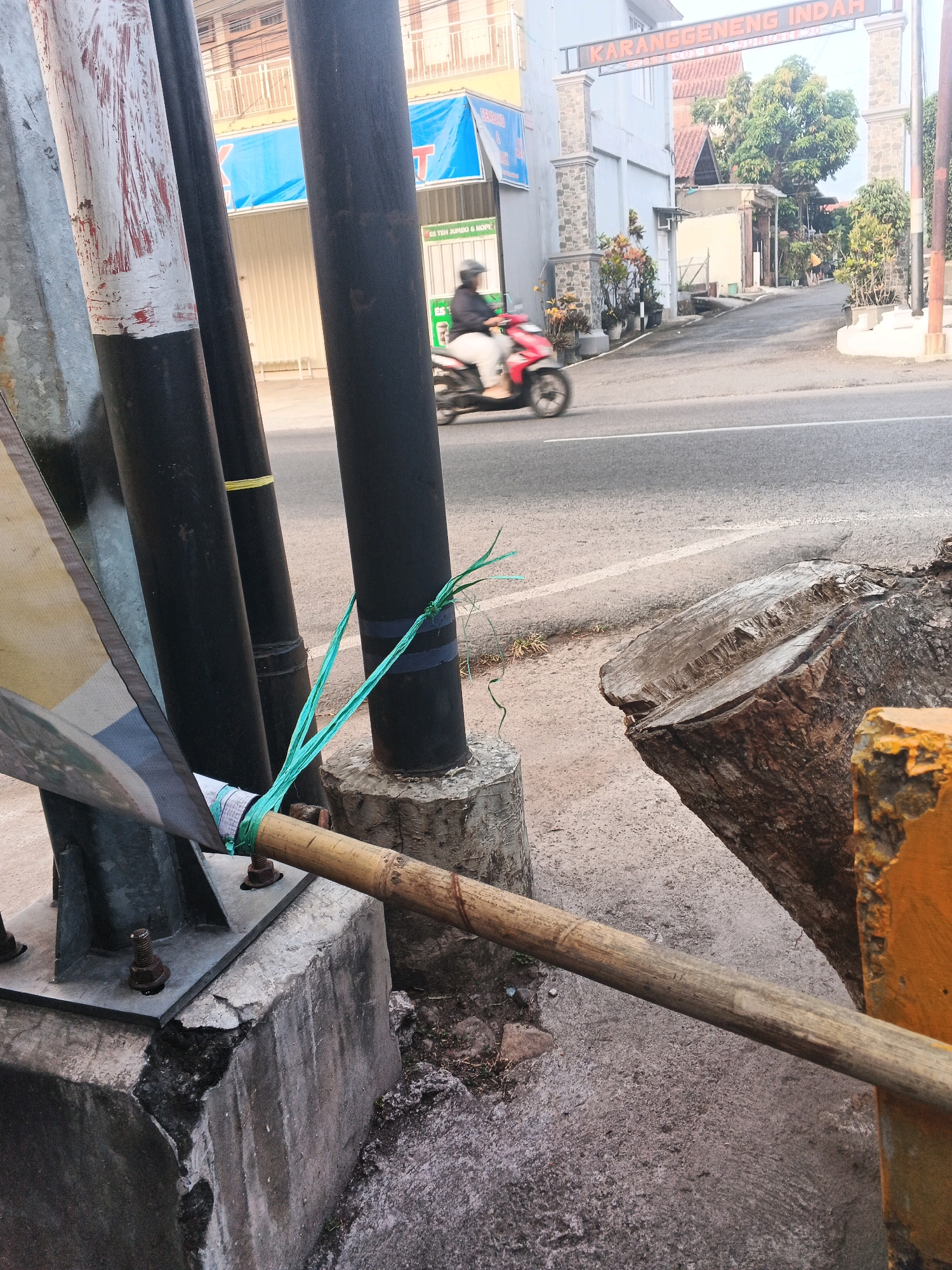 In Semarang, Indonesia, a close-up, eye-level shot captures a section of a road and its surrounding elements. A motorbike, ridden by a person wearing a helmet and white pants, is captured in motion, blurring across the asphalt. The road is divided by a white line and shows signs of wear. To the left, three dark posts stand vertically, one of which is partially covered by a yellow and black striped fabric. A bamboo pole is secured to one of these posts with green string. In the foreground, a large tree stump, freshly cut, rests on the ground. Behind the stump, a yellow painted object is visible. The background features buildings with signs, including one that reads "KARANGGENG INDAH," and lush green trees. The scene is bathed in natural light, indicating it was taken during the day.