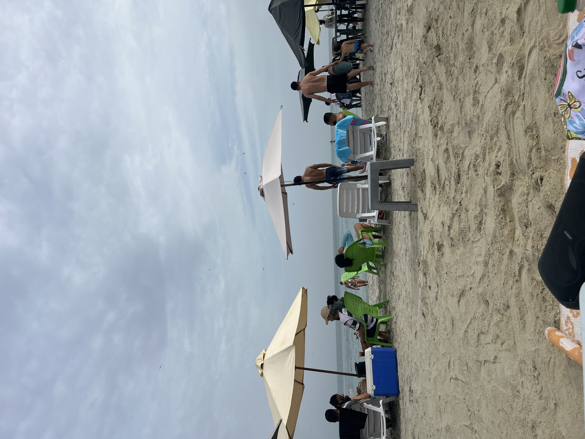 The scene is set on a beach in Porlamar, Venezuela. The weather is overcast. Several groups of people are relaxing on the sand. There are white and green plastic chairs set up, and beach umbrellas are in place to provide shade. People are seen lounging in the chairs, while others are walking. In the foreground, there is a portion of a chair and a person's leg.