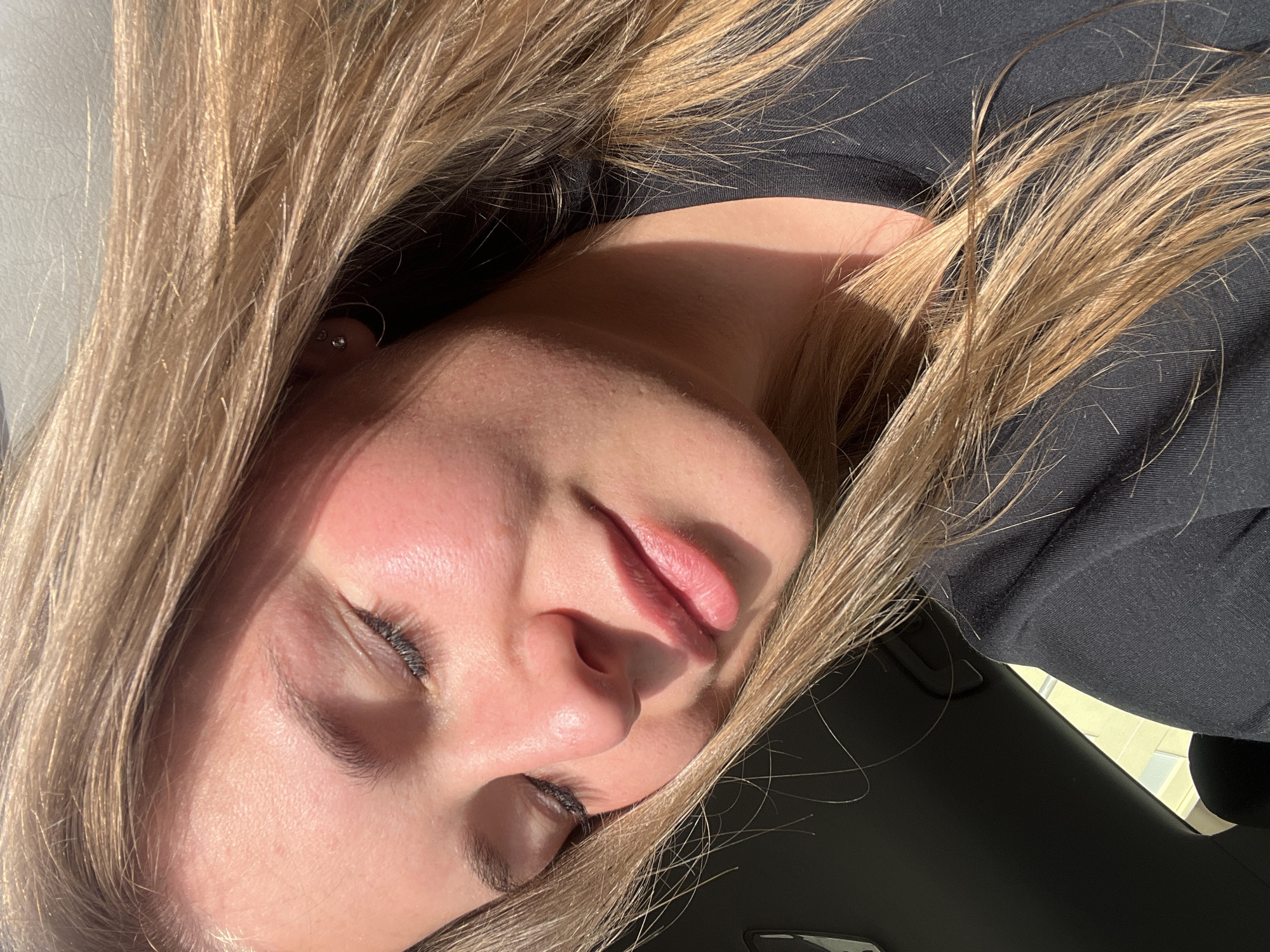 The image shows a woman with blonde hair and closed eyes, lying down in a location suggestive of a car. She is wearing a dark-colored top. It is not possible to determine the location of Budva, Montenegro from the given image.